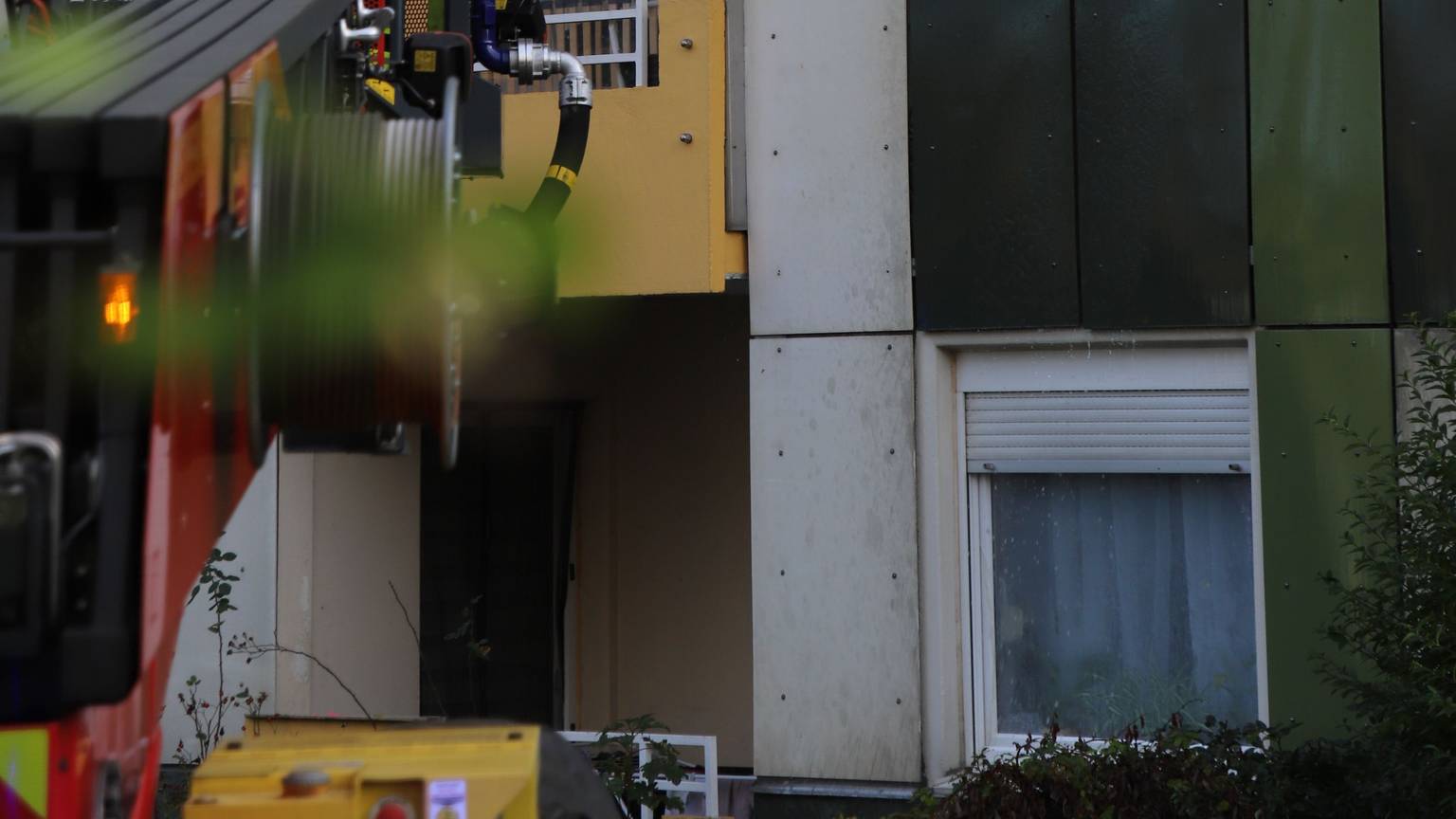 Ein Feuerwehrmann steht auf einer Drehleiter und löscht mit einem Schlauch das Feuer hinter einem Fenster. Es steigt schwarzer Rauch auf.