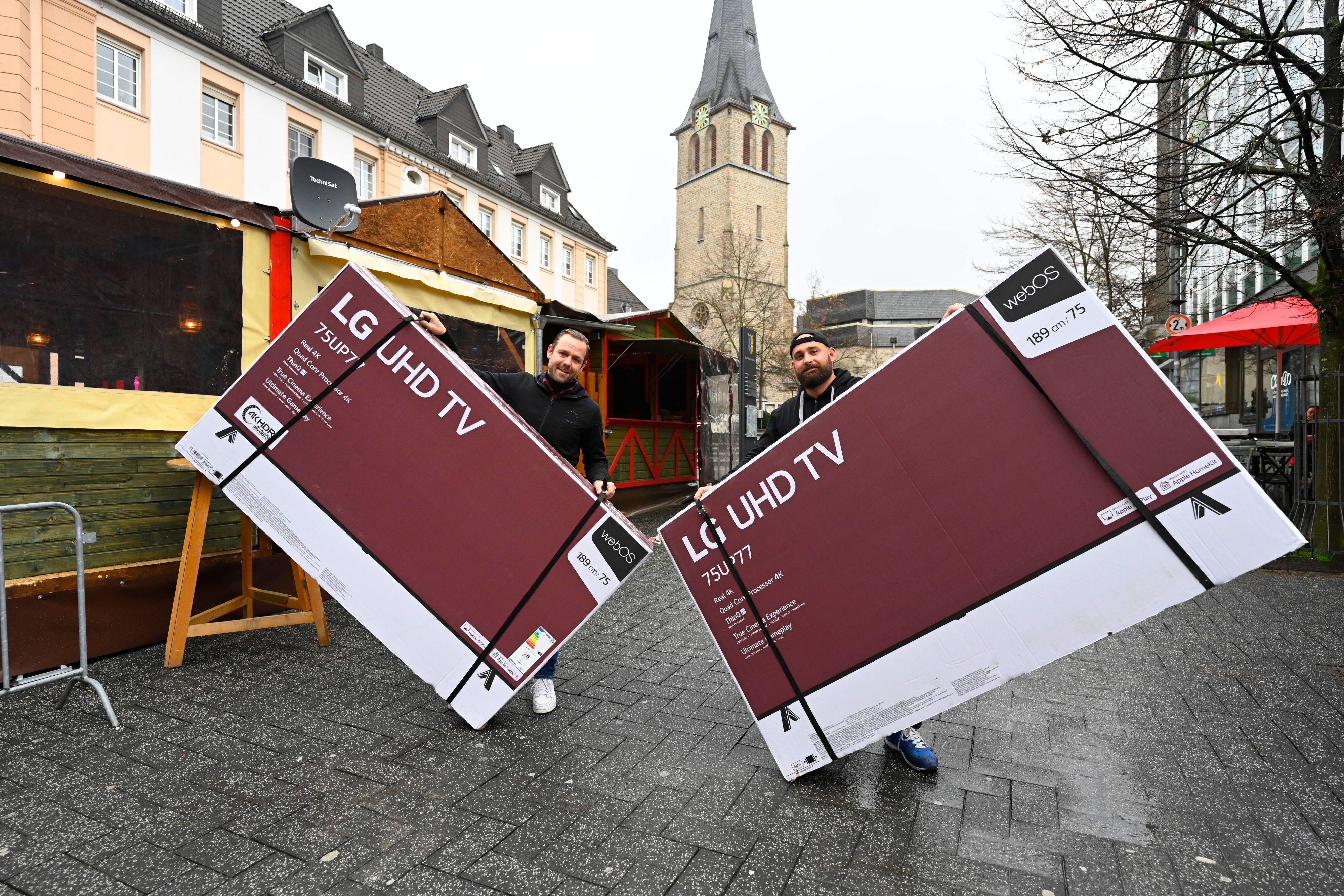 Der Gastronom Timo Bay hält einen großen verpackten Bildschirm in der Hand, ebenso sein Freund. Sie stehen in der Gummersbacher Innenstadt vor einem Glühweinstand und zeigen hier ab Sonntag die Fußball-WM.