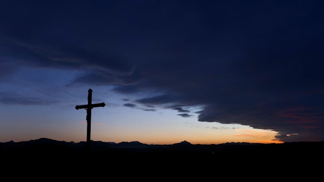 Eine Wolkenfront zieht hinter einem Wegkreuz über die im Sonnenuntergang liegende Berglandschaft.