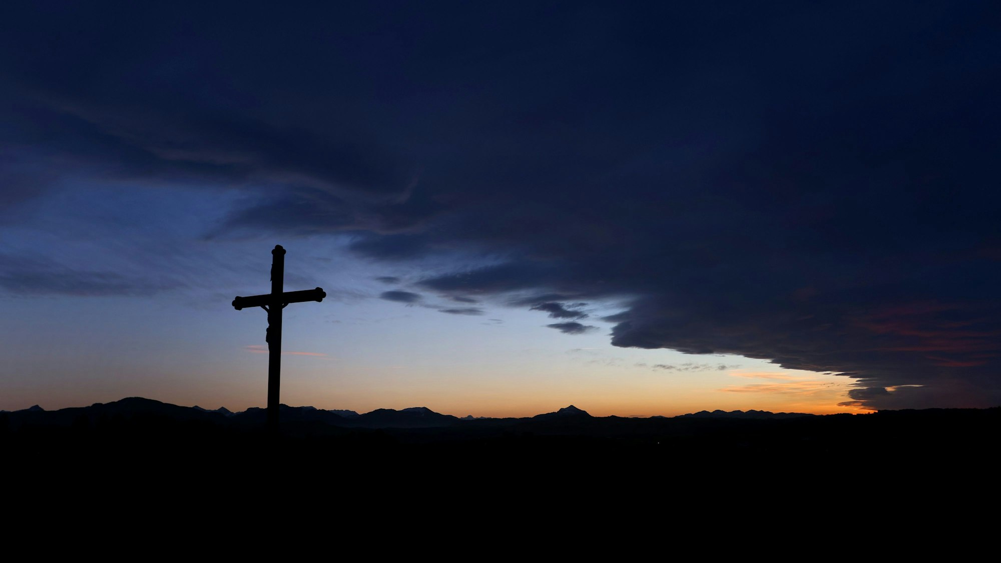 Eine Wolkenfront zieht hinter einem Wegkreuz über die im Sonnenuntergang liegende Berglandschaft.