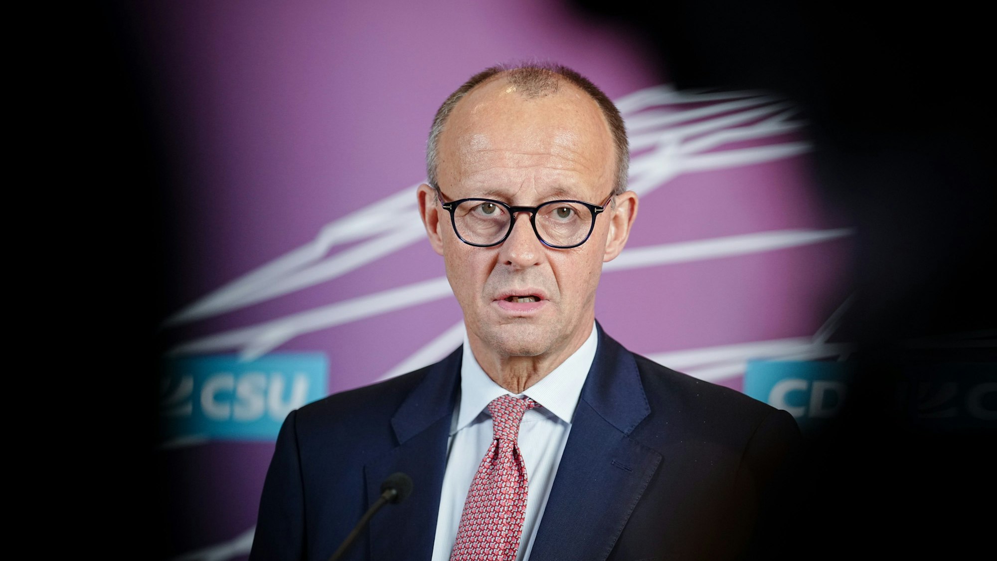 Merz steht vor einer Wand mit Unionslogo, redet und guckt in die Kamera. Am Rand des Bildes sieht man schwarze Schatten.