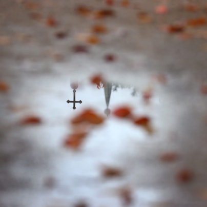 Ein Kreuz und eine Kirchturmspitze spiegelt sich in einer Wasserlache.