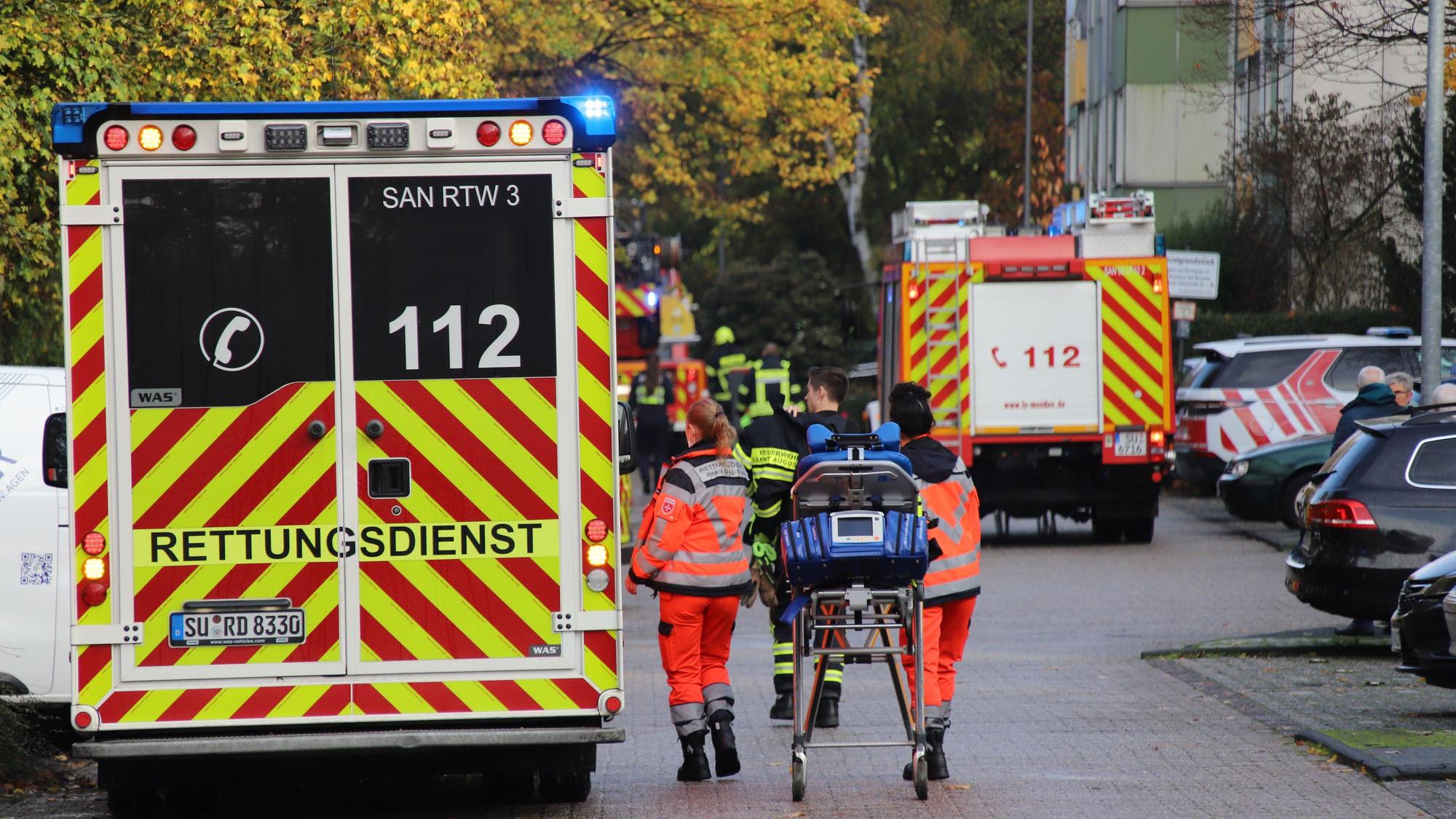 Einsatzkräfte gehen mit einer Liege zu einem Rettungswagen. Im Hintergrund steht ein Feuerwehrauto und mehrere Feuerwehrleute.