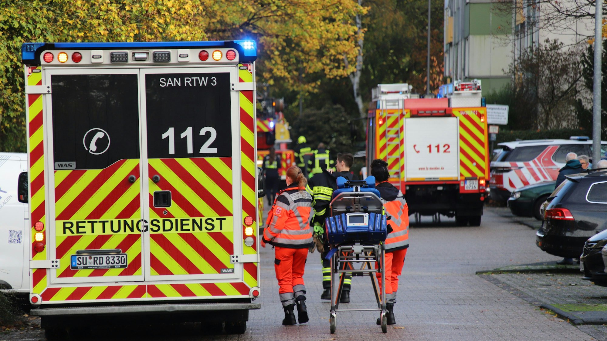 Einsatzkräfte gehen mit einer Liege zu einem Rettungswagen. Im Hintergrund steht ein Feuerwehrauto und mehrere Feuerwehrleute.