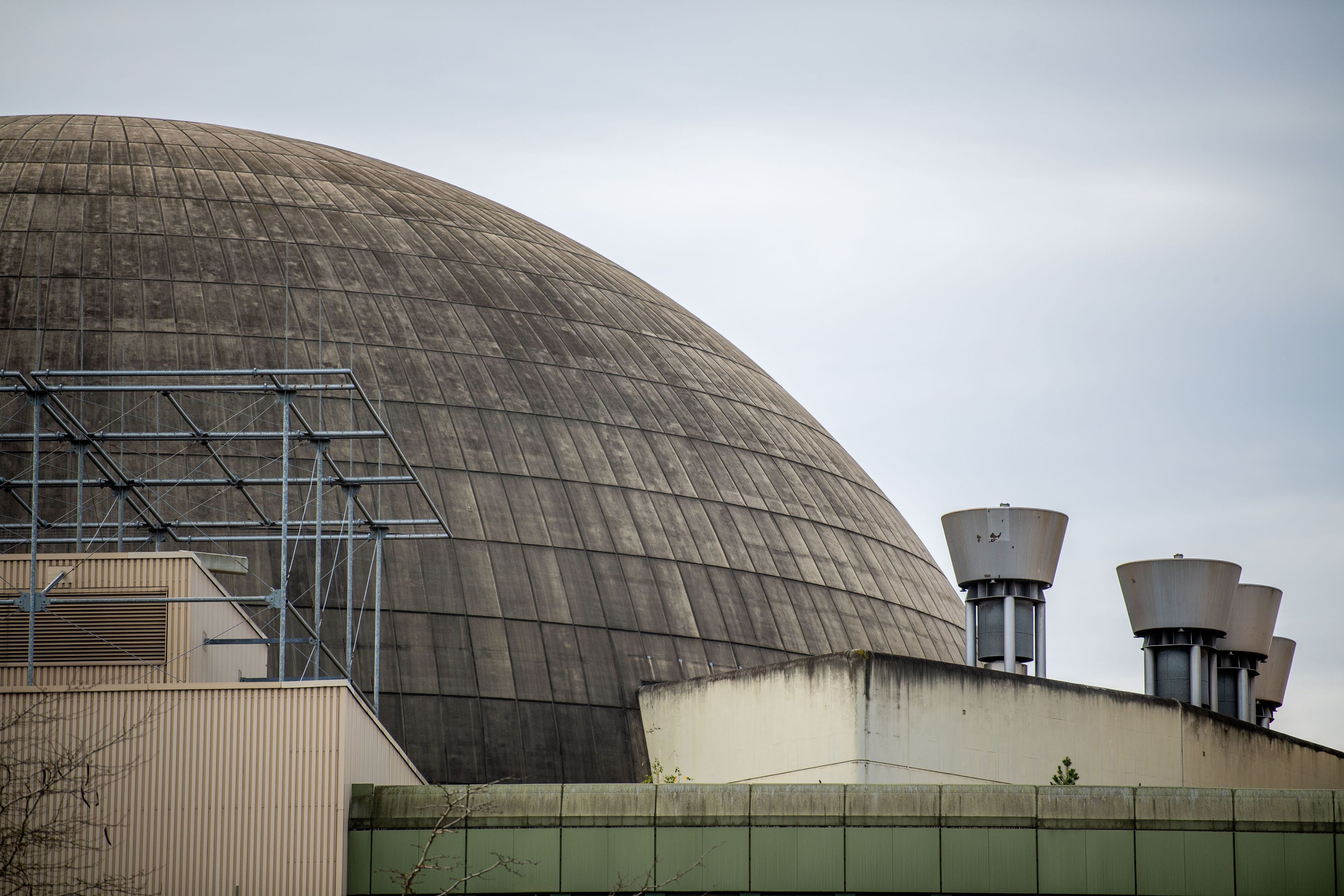 Man sieht den rechten Teil einer Betonhalbkugel. Im Vordergrund der obere Teil einer Industriehalle, links ein Gerüst auf dem Dach, rechts Schornsteine. Im Hintergrund sieht man den Himmel.