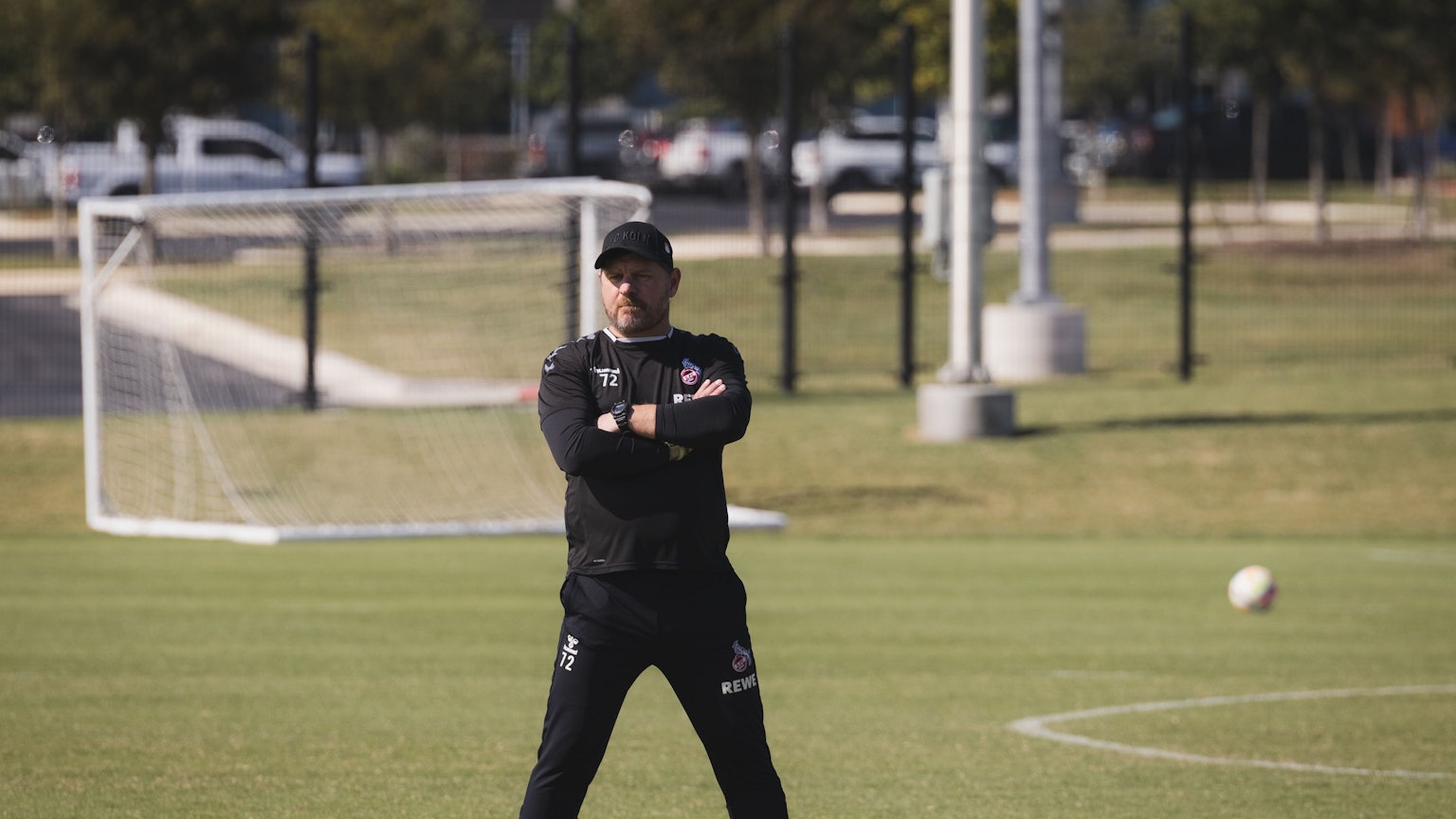 Steffen Baumgart steht breitbeinig und mit verschränkten Armen auf dem Spielfeld. Er trägt schwarze Sportkleidung und eine Kappe. Im Hintergrund sieht man ein Tor und einen Fußball auf dem Feld liegen.