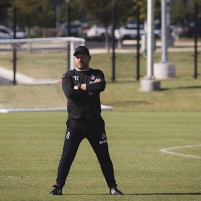 Steffen Baumgart steht breitbeinig und mit verschränkten Armen auf dem Spielfeld. Er trägt schwarze Sportkleidung und eine Kappe. Im Hintergrund sieht man ein Tor und einen Fußball auf dem Feld liegen.