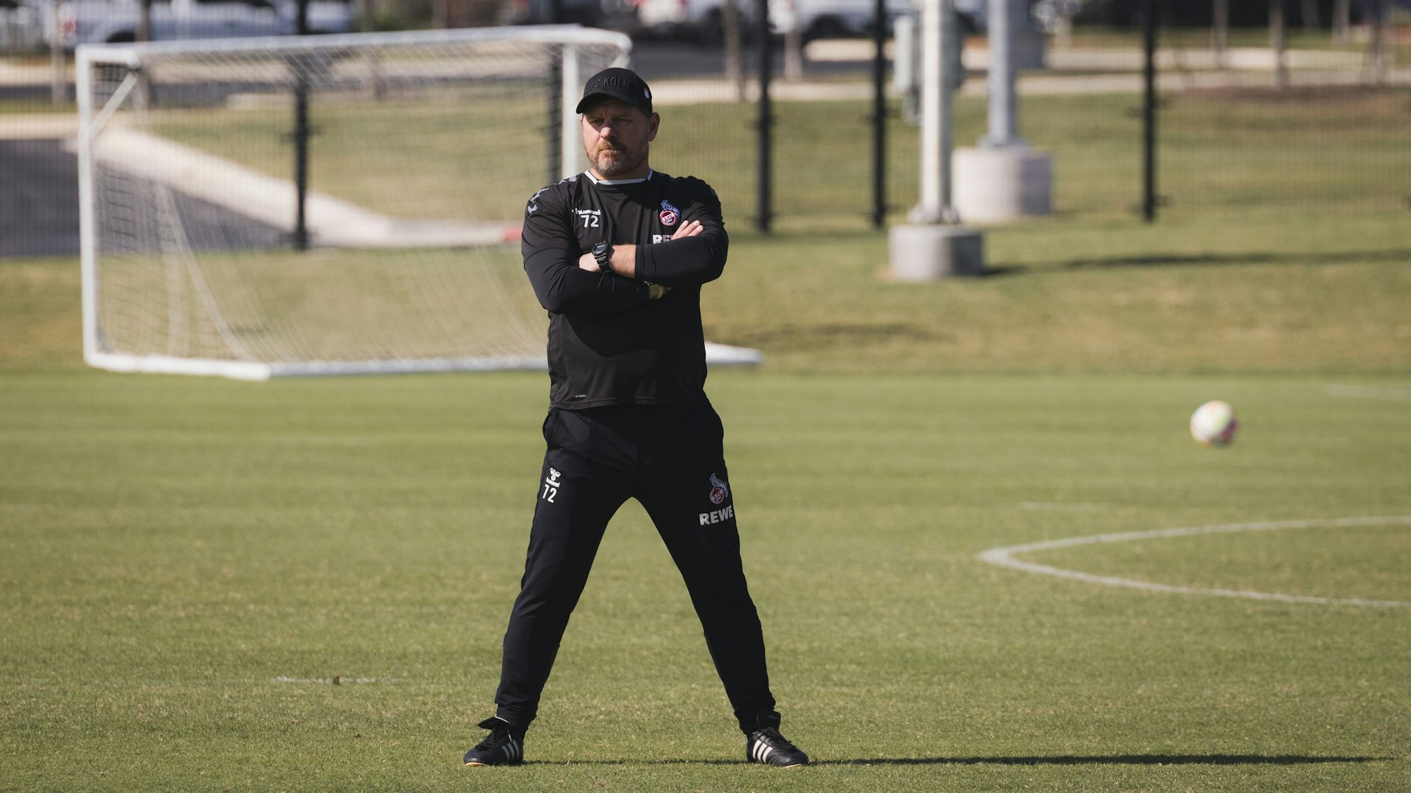 Steffen Baumgart steht breitbeinig und mit verschränkten Armen auf dem Spielfeld. Er trägt schwarze Sportkleidung und eine Kappe. Im Hintergrund sieht man ein Tor und einen Fußball auf dem Feld liegen.