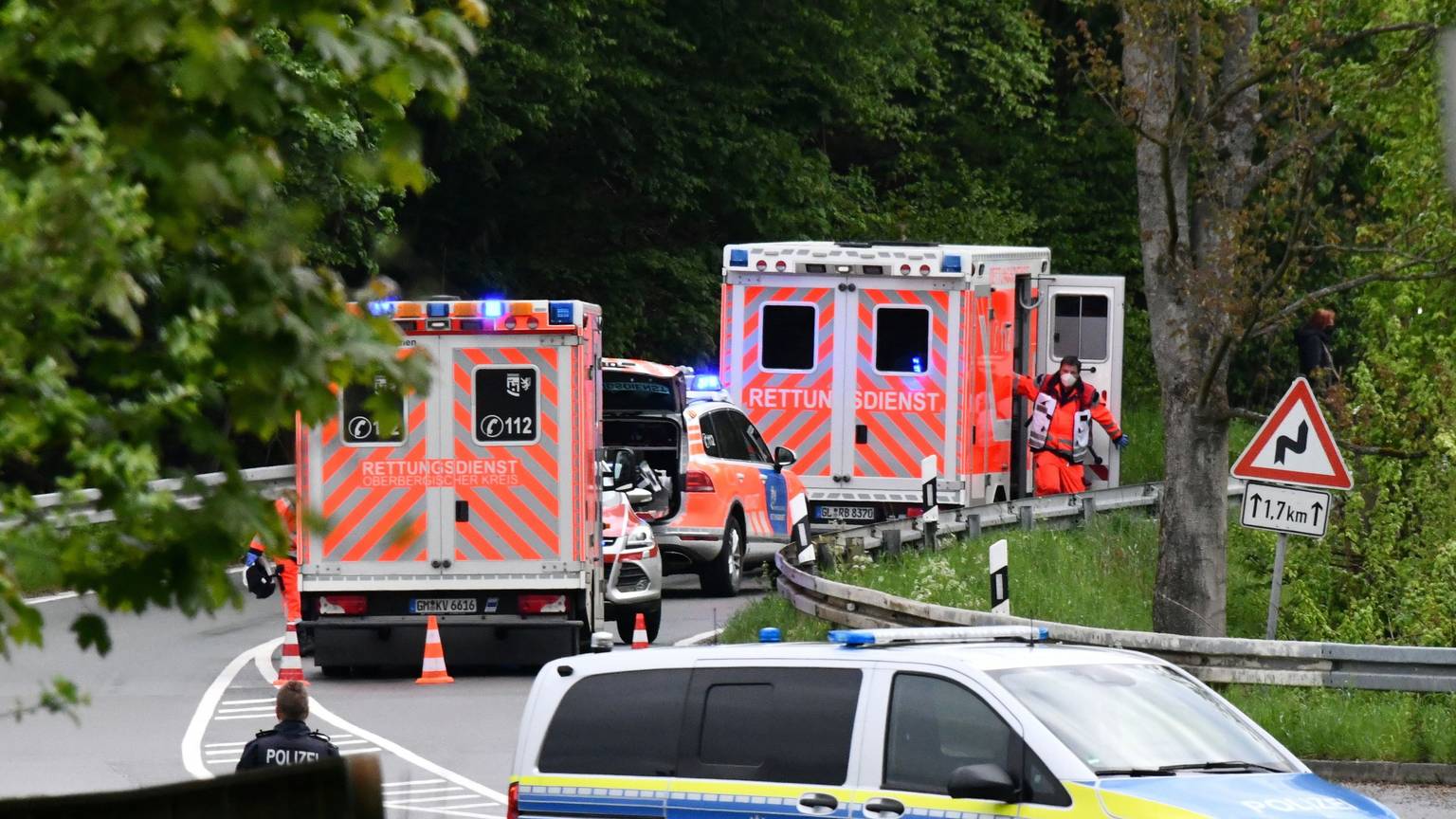Im Vordergrund steht ein Polizeiwagen quer auf der Straße, weiter hinten sieht man zwei Krankenwagen und einen Notarztwagen am Straßenrand stehen. Die Straße führt durch einen Wald.