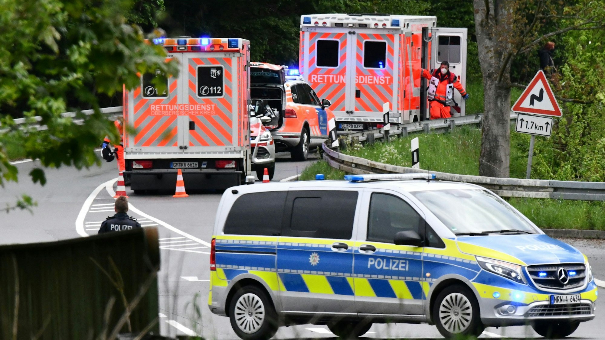 Im Vordergrund steht ein Polizeiwagen quer auf der Straße, weiter hinten sieht man zwei Krankenwagen und einen Notarztwagen am Straßenrand stehen. Die Straße führt durch einen Wald.