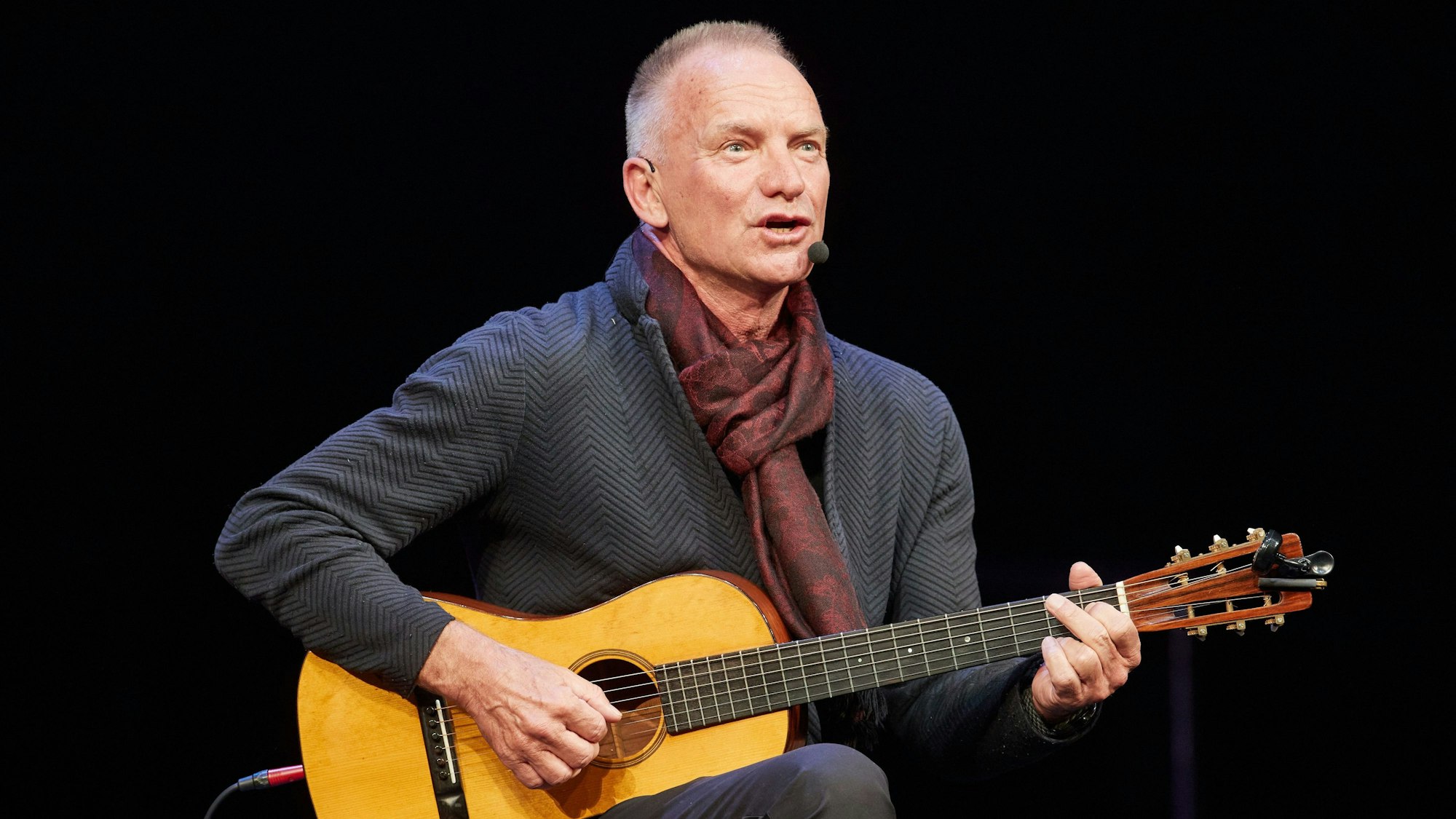 Zu sehen ist Sting, wie er auf der Bühne sitzt, er hält eine Gitarre in den Händen und singt.