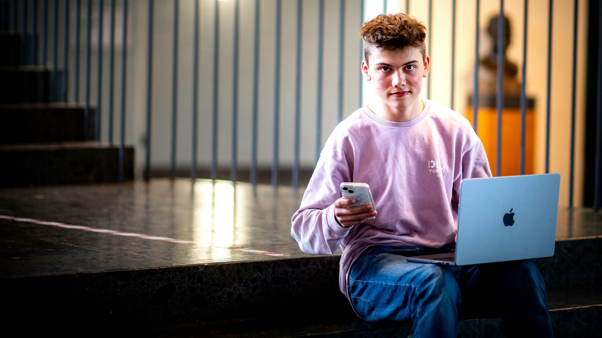 Handy in der Hand, Laptop auf den Beinen. Schüler Noel Dederichs sitzt auf den Stufen des Emil-Fischer-Gymnasiums in Euskirchen.