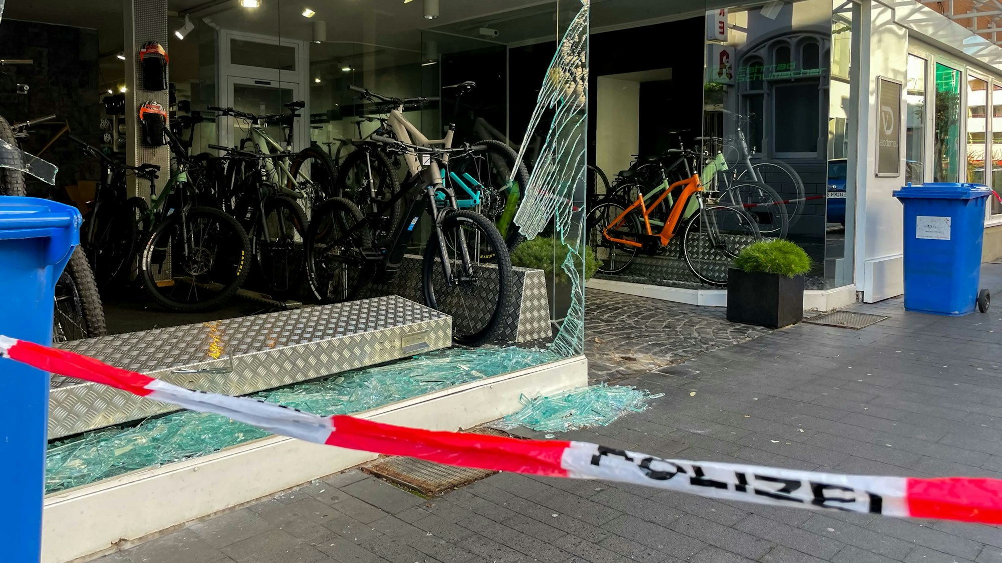 Das zertrümmerte Schaufenster eines Fahrradladens. Auf dem Boden und in der Auslage liegen Glasscherben, einige Fahrräder sind noch ausgestellt. Im Vordergrund steht auf weiß-rotem Absperrband das Wort Polizei.
