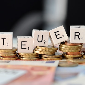 Würfel mit den Buchstaben S t e u e r liegen auf Geldmünzen und Geldscheinen.