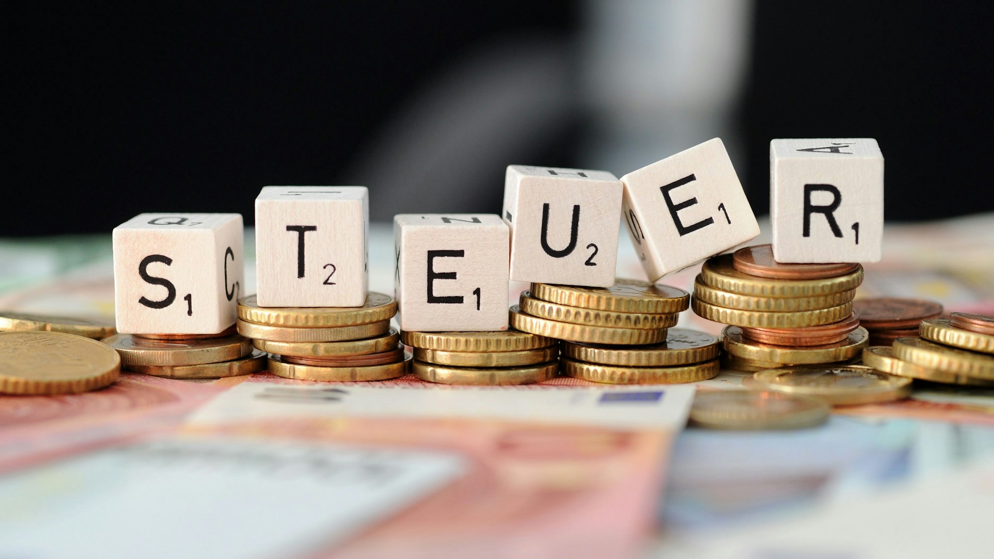 Würfel mit den Buchstaben S t e u e r liegen auf Geldmünzen und Geldscheinen.