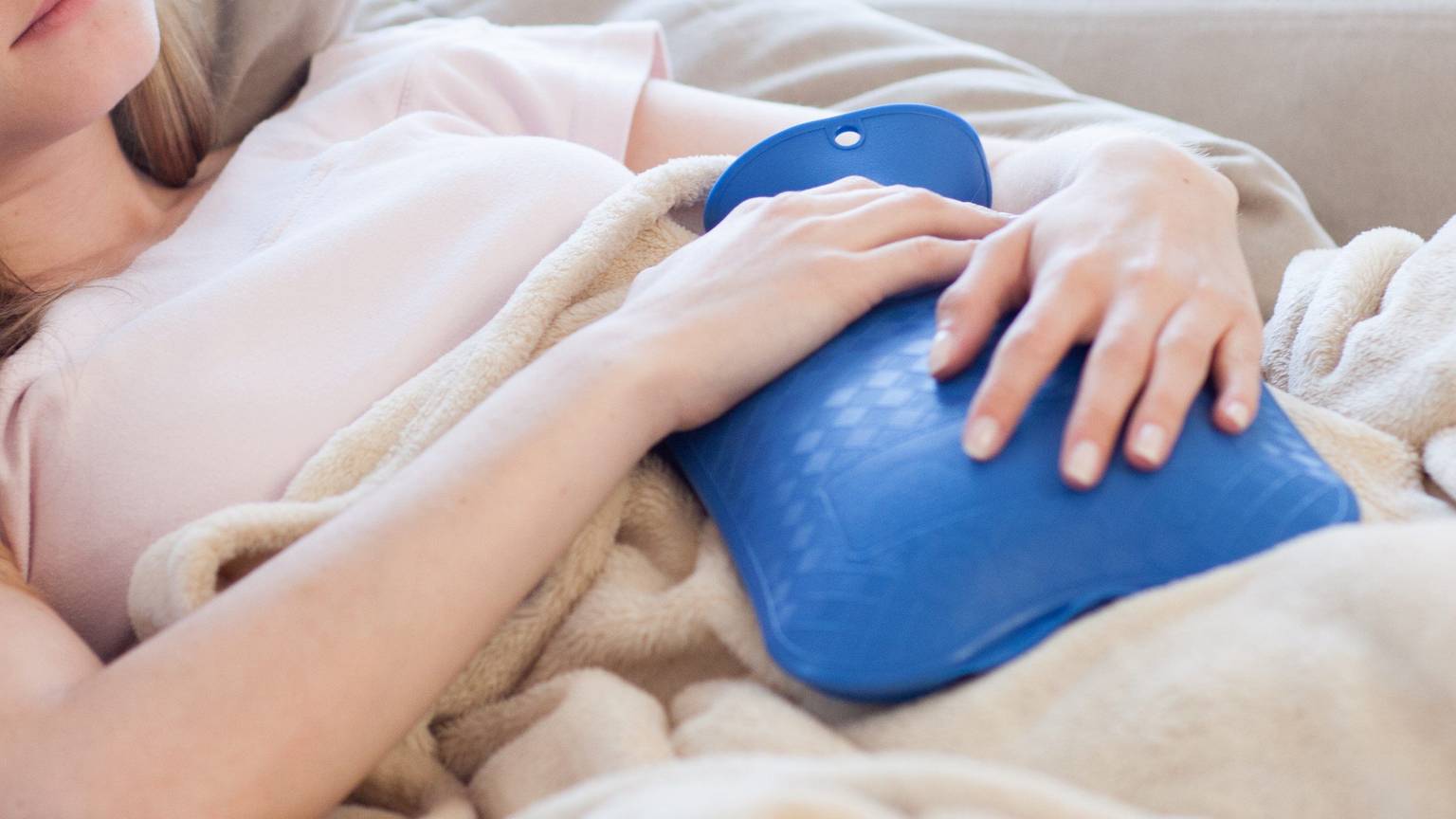 Eine Frau liegt mit einer blauen Wärmflasche auf dem Bauch mit einer Decke auf dem Sofa.