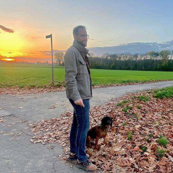 Ein Mann blickt auf ein Feld, sein Hund sitzt neben ihm.