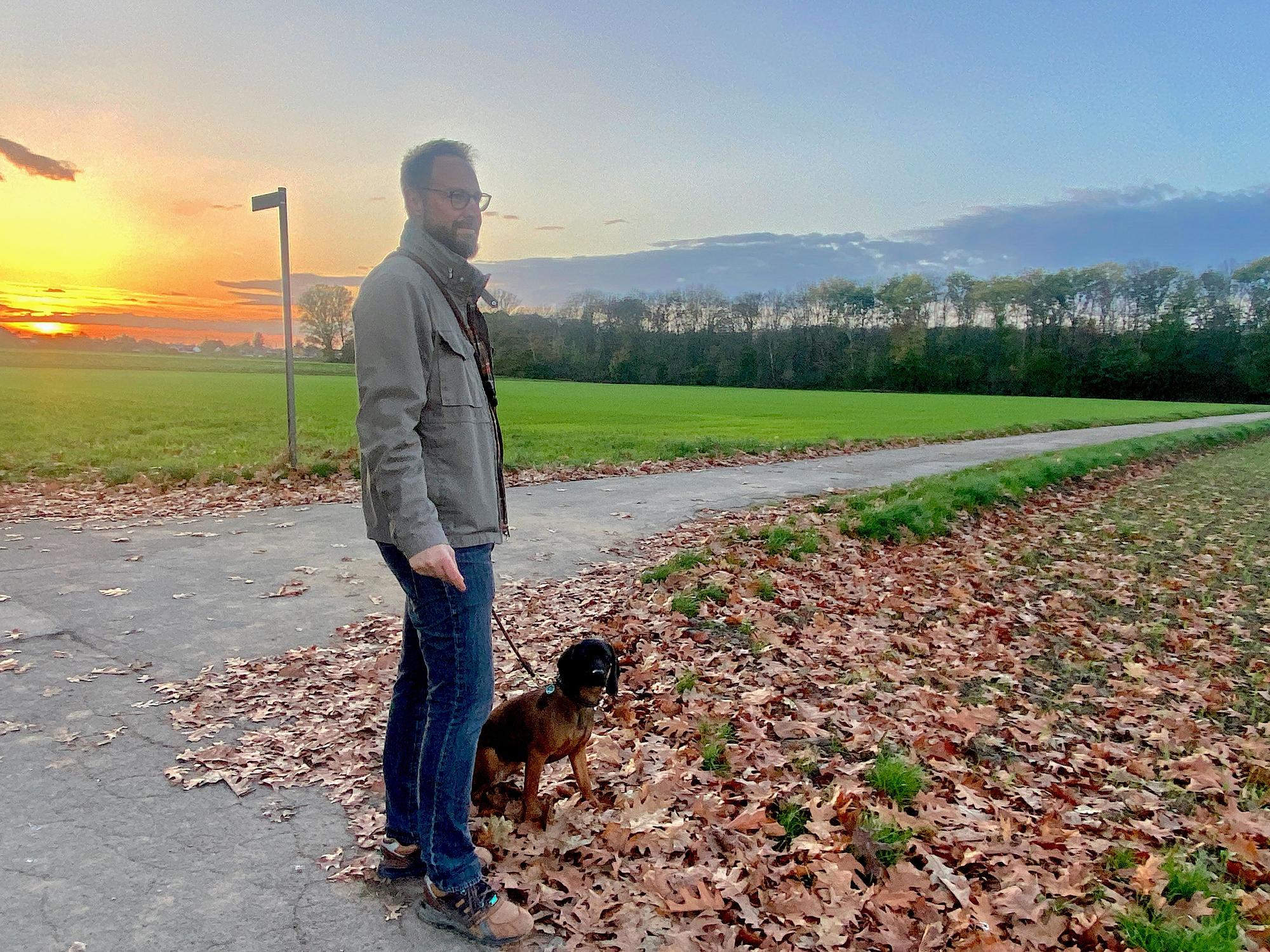 Ein Mann blickt auf ein Feld, sein Hund sitzt neben ihm.