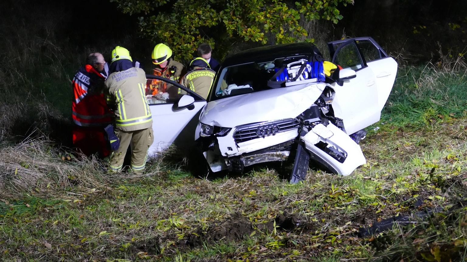 Ein weißes Auto steht im Graben, die Türen stehen offen, das Heck ist demoliert. Daneben stehen Feuerwehrleute und ein Notarzt.