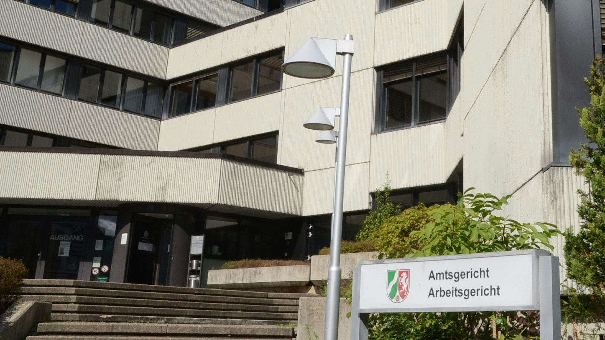 Der Eingang des Amtsgerichts in Siegburg. Vor dem Gebäude steht ein Schild mit dem nordrheinwestfälischen Wappen und der Aufschrift „Amtsgericht/Arbeitsgericht“.