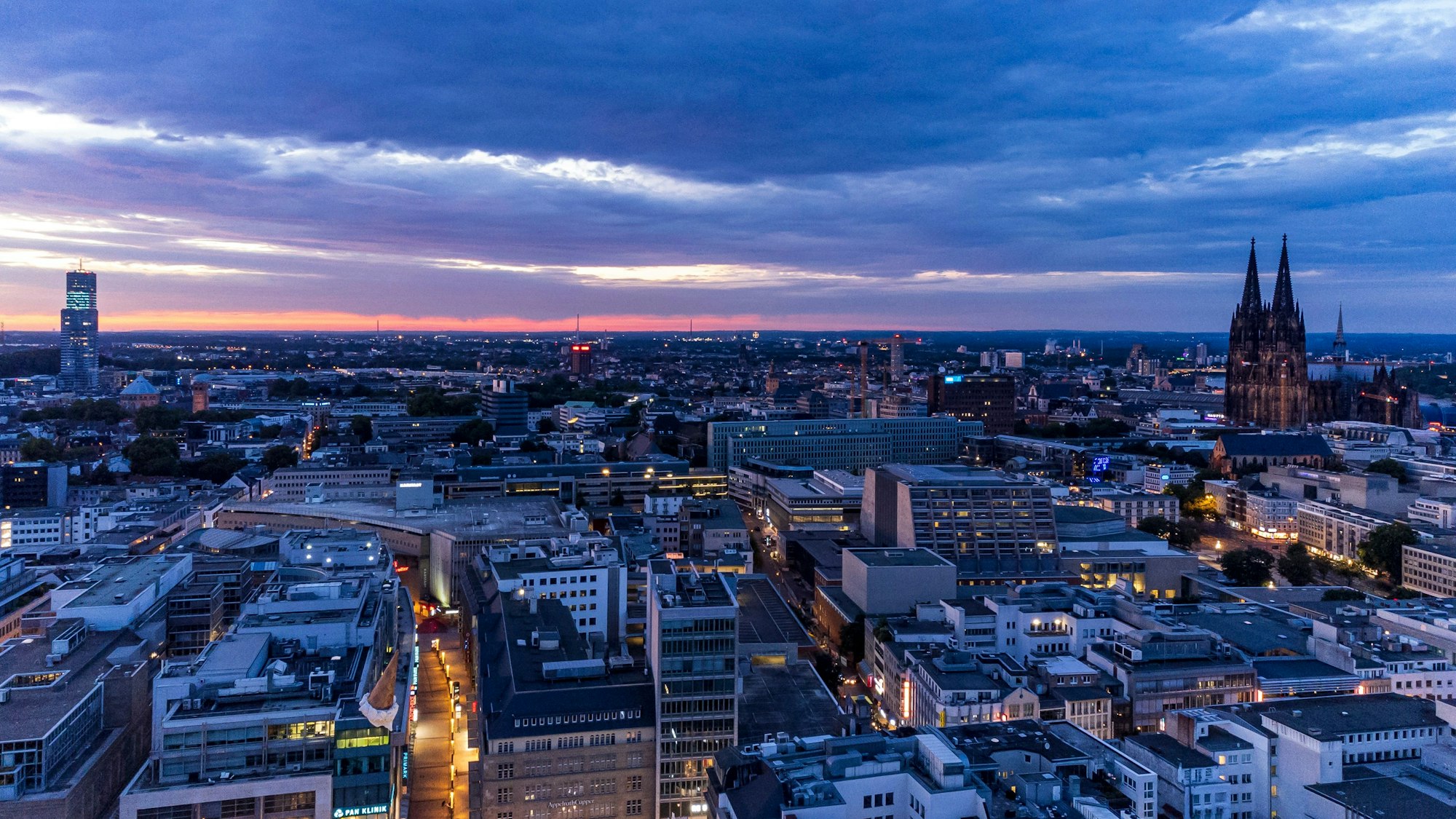 Das Foto ist über die Kölner Altstadt fotografiert, links der Dom. Der Himmel ist wolkig, die Sonne geht unter.