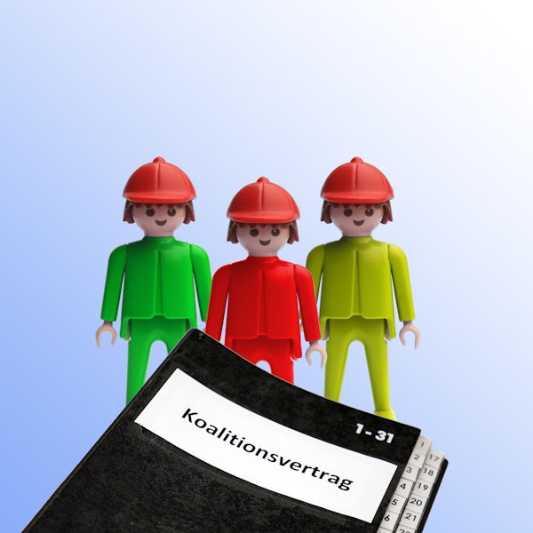 Ein grüner, ein roter und ein gelber Playmobilmensch stehen hinter einer Mappe mit der Aufschrift „Koalitionsvertrag“.