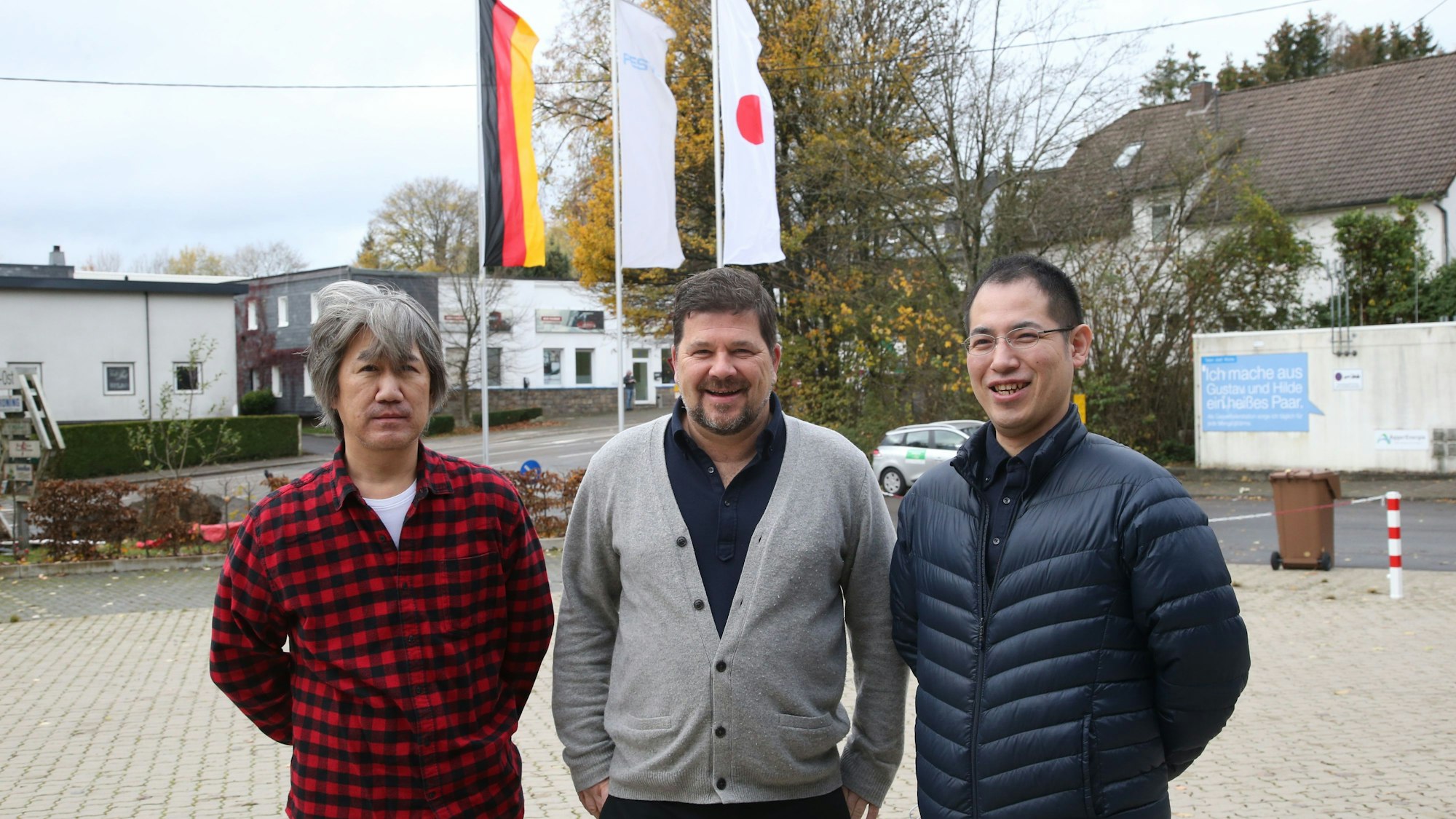Drei Männer stehen auf einem Parkplatz, im Hintergrund sind die deutsche und die japanisache Fahne zu sehen.
