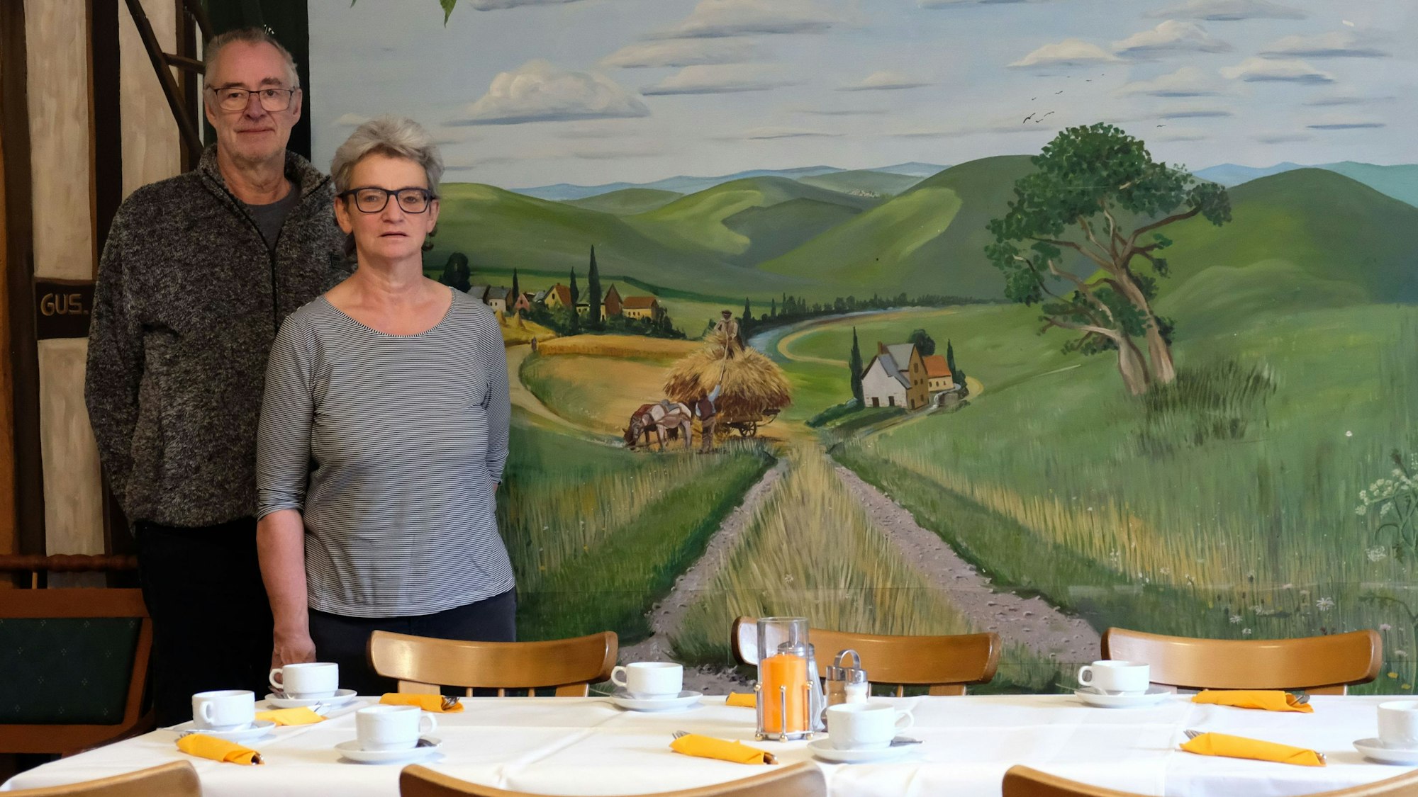 Annegret und Volker Prause, die seit 2004 den "Brunnenhof" in Mechernich-Holzheim betrieben haben, stehen vor einem Tisch, hinter ihnen ist ein Feldweg auf eine Wand gemalt.