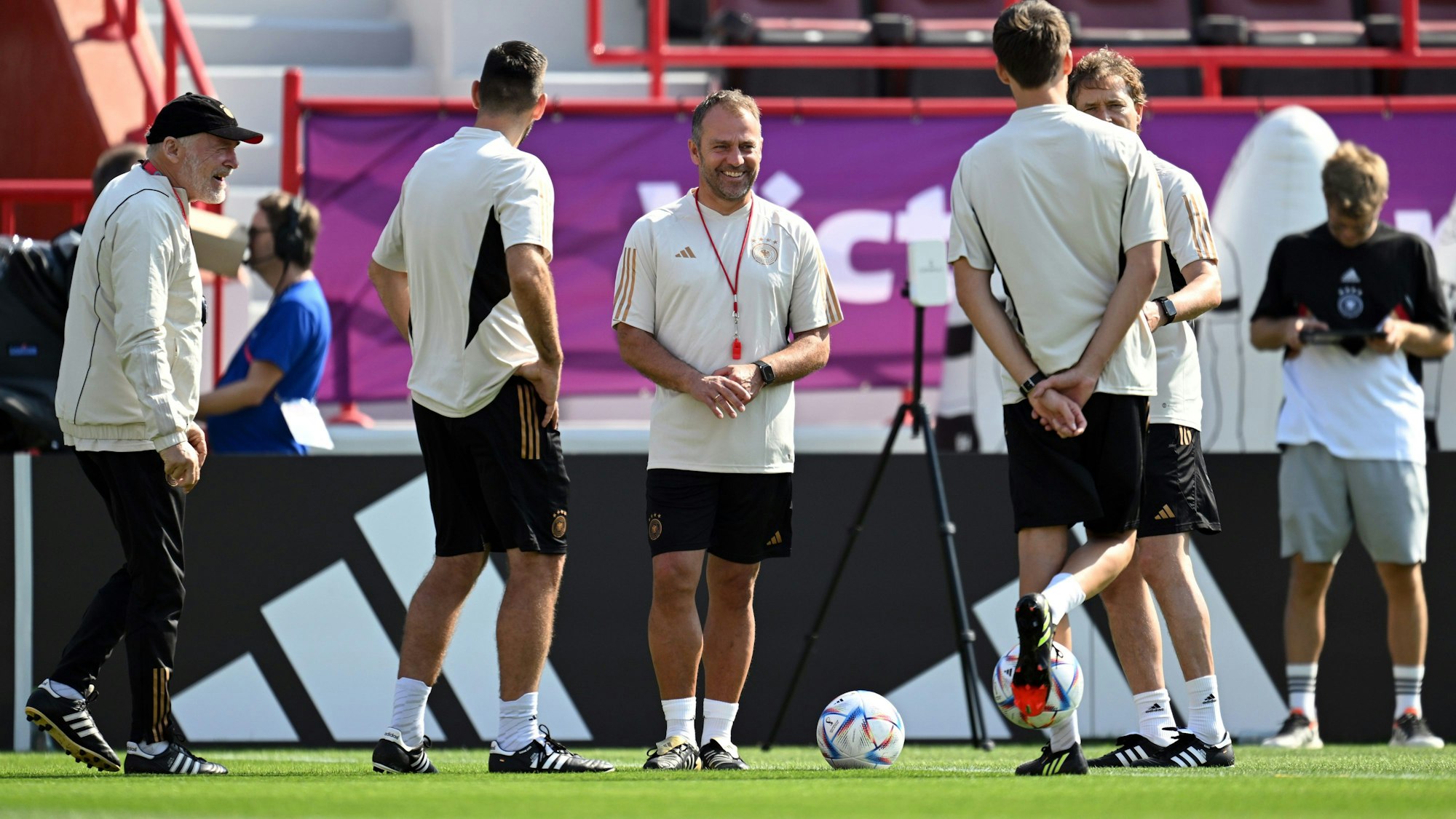 Hansi Flick und sein Trainerteam stehen auf einem Fußballfeld, sie sind gut gelaunt, ein Ball liegt in der Mitte der Gruppe.