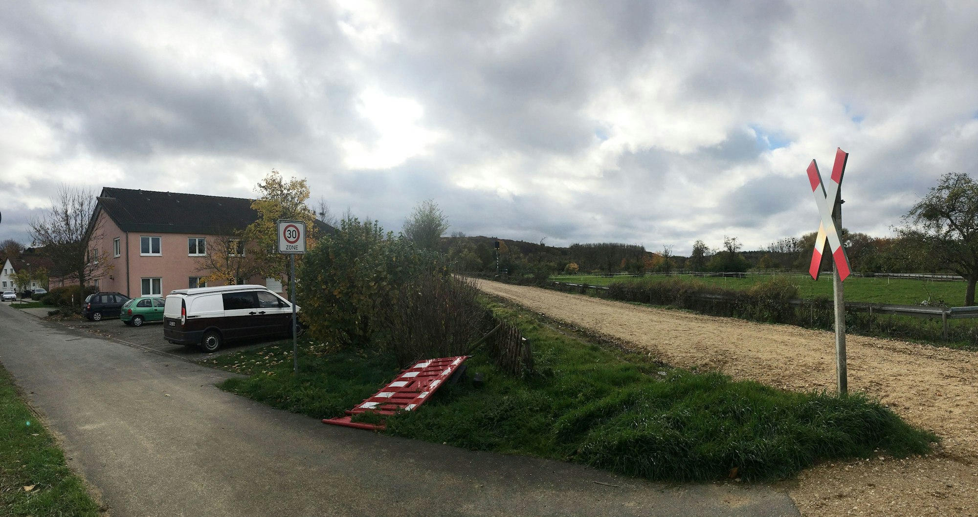 Auf der Bahnstrecke liegen keine Gleise. Neben der Strecke liegt ein Haus. Davor stehen Autos.