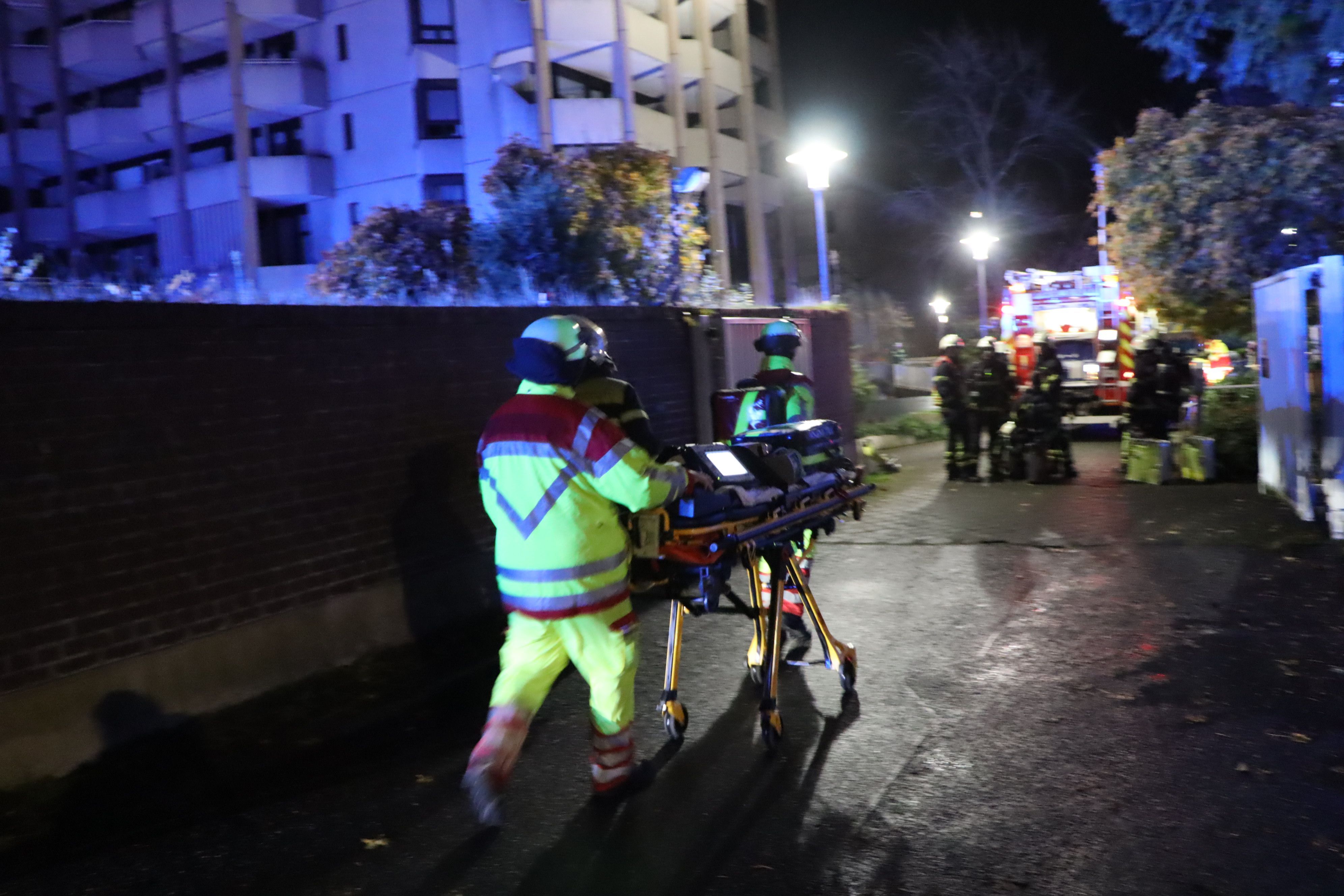 Einsatzkräfte der Feuerwehr schieben eine Trage über die Straße. Es ist Nacht, die Szene ist von Blaulicht erleuchtet.