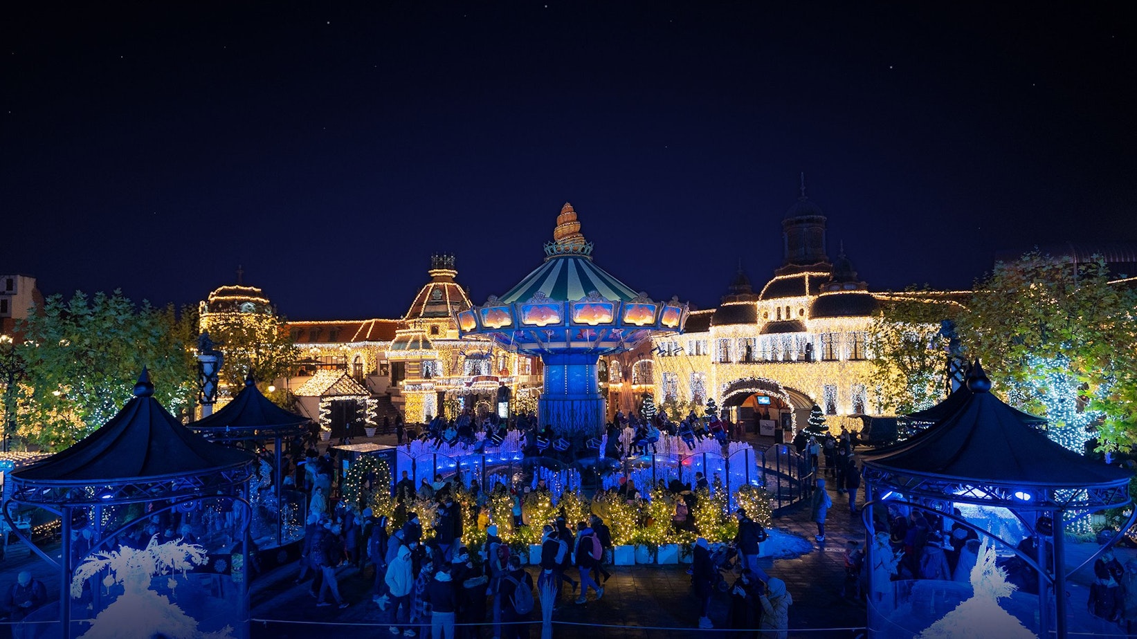 Die Gebäude im Hintergrund sind mit vielen Lichtern beleuchtet, das Kettenkarussell leuchtet blau, viele Besucher stehen davor.