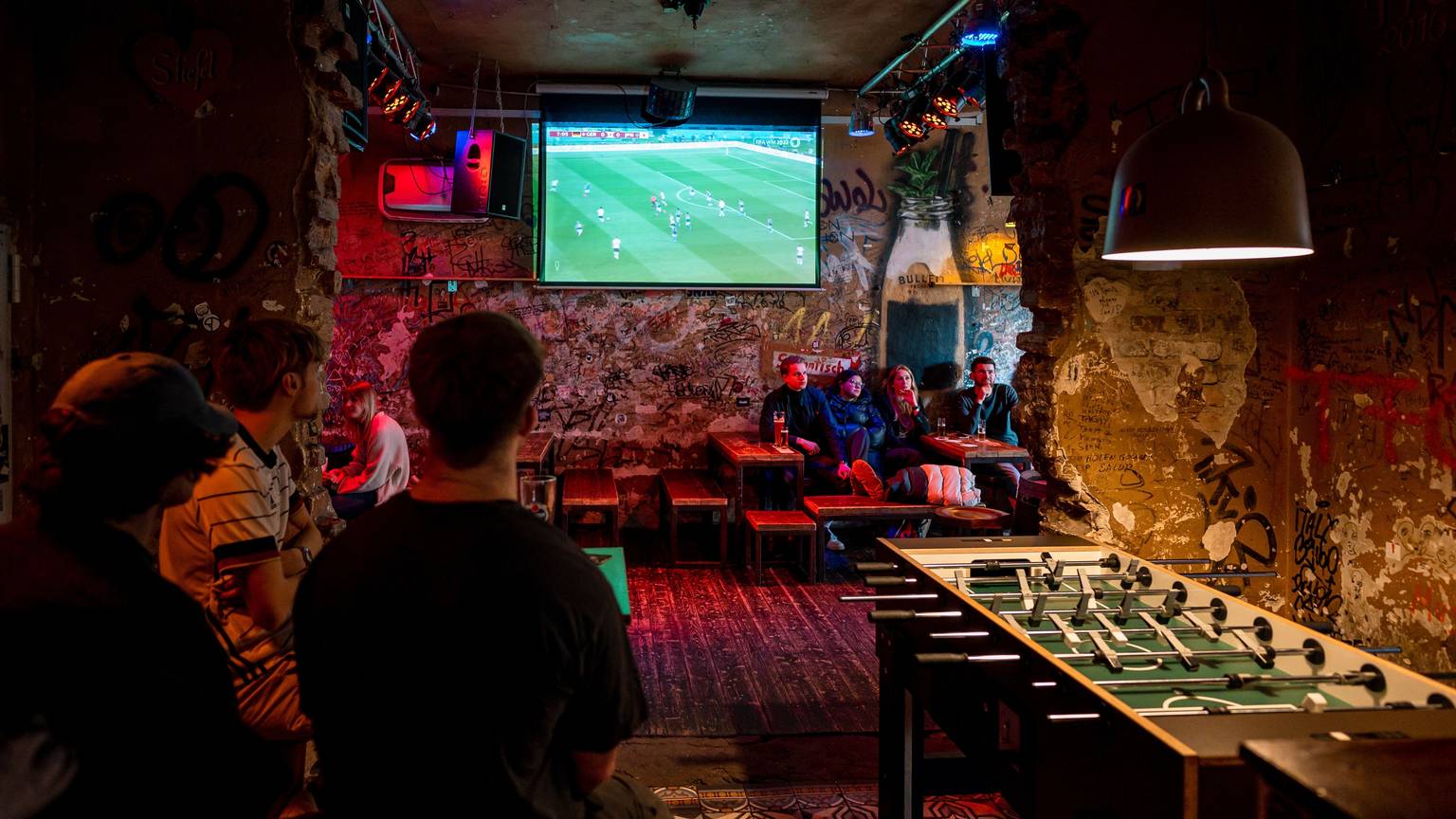 Im „Stiefel“ auf der Zülpicher Straße verfolgten einige wenige Kölner das WM-Auftaktspiel der deutschen Mannschaft gegen Japan.Das Bild zeigt drei junge Männer an einem Tisch, im Hintergrund hängt der TV-Bildschirm.