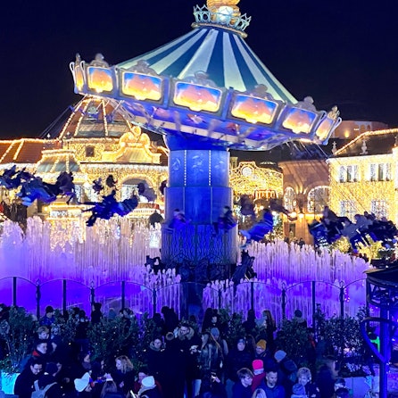 Zu sehen ist ein in bunten Farben beleuchtetes Kettenkarussell im Phantasialand sowie mit Lichter geschmückte Häuser im Hintergrund.