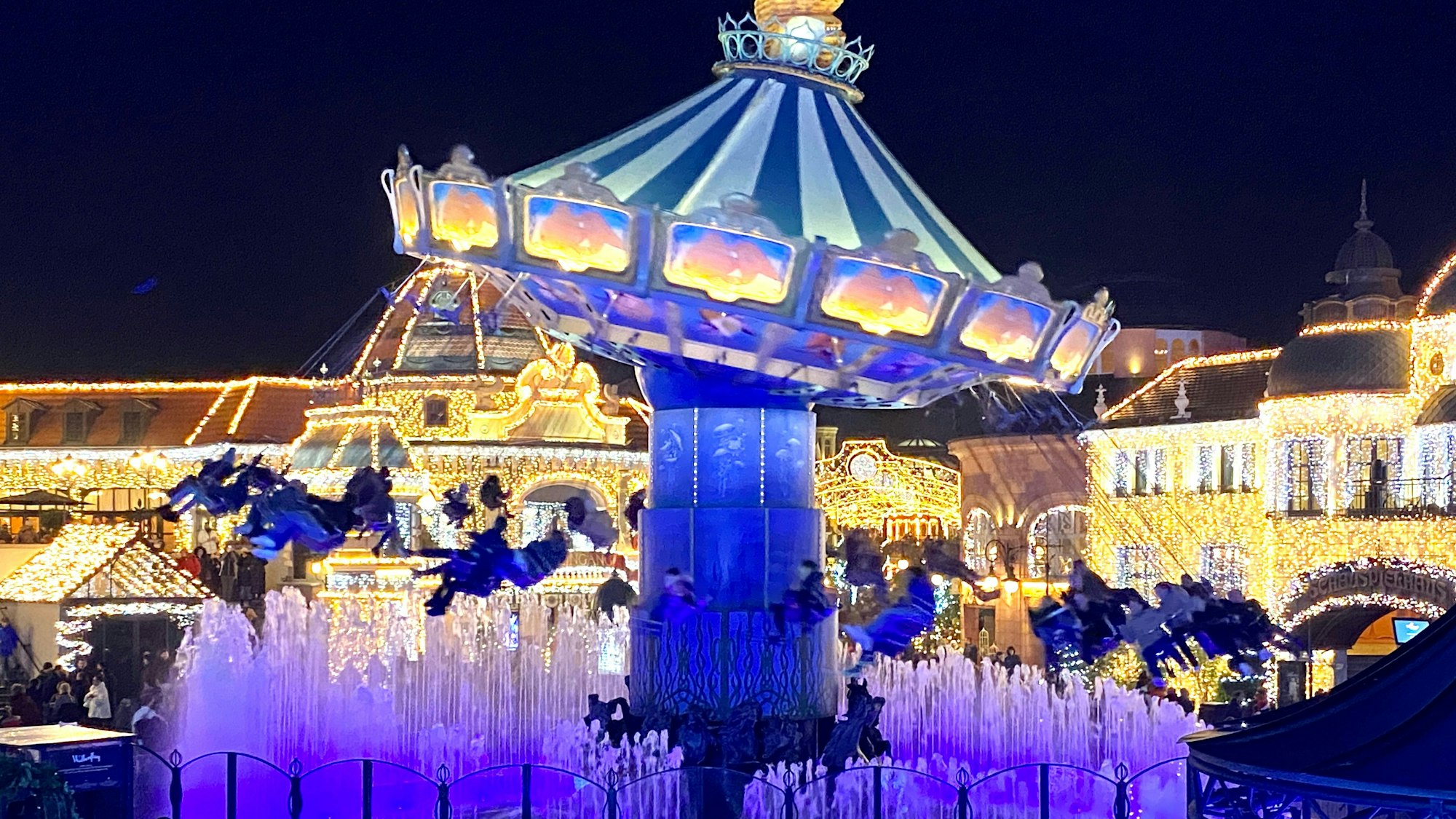 Zu sehen ist ein in bunten Farben beleuchtetes Kettenkarussell im Phantasialand sowie mit Lichter geschmückte Häuser im Hintergrund.