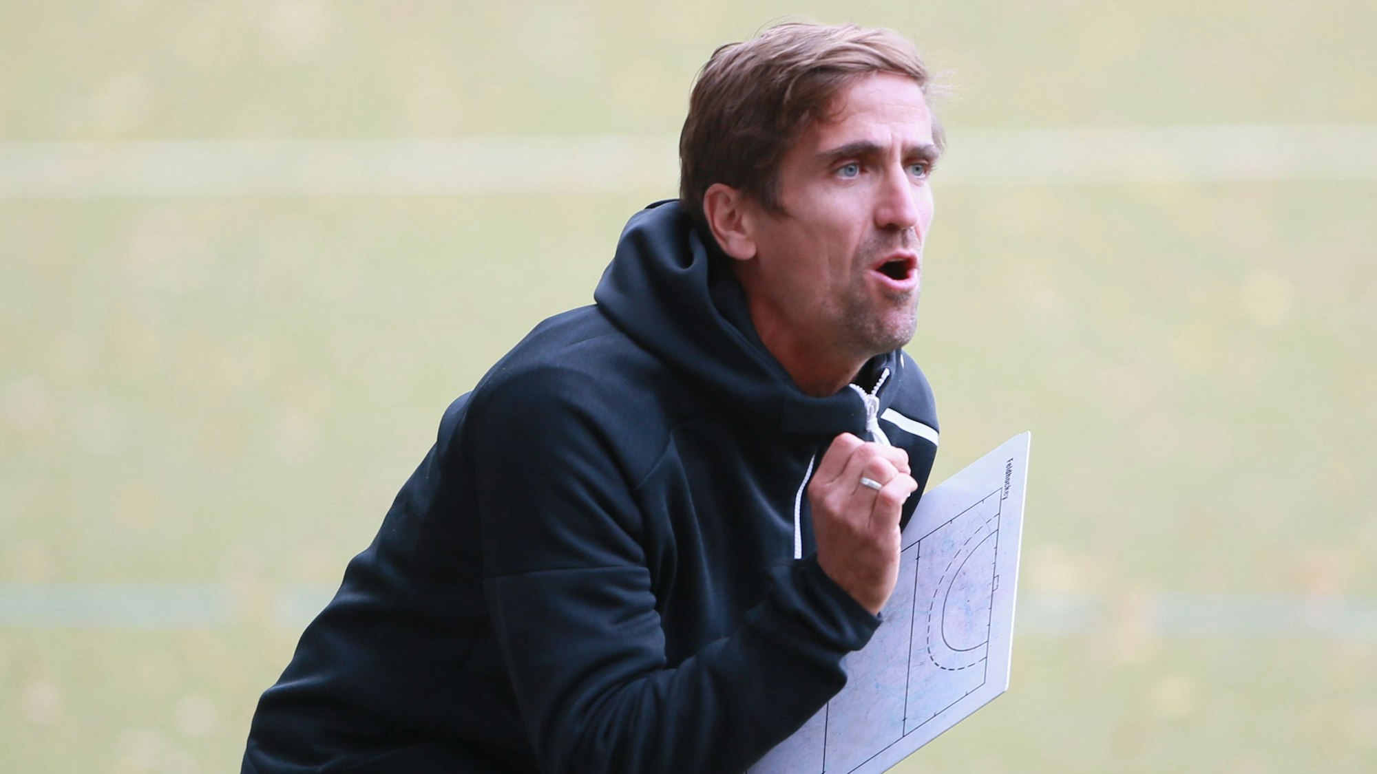 Ein Hockeytrainer steht mit seinem Taktikbrett am Spielfeldrand und feuert seine Mannschaft an.
