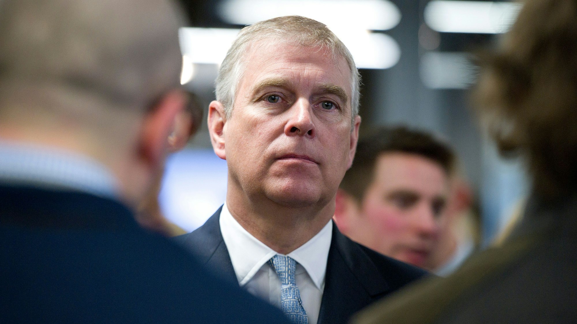 Prinz Andrew, Herzog von York, schaut bei einem Gespräch ernst in die Kamera. Er trägt einen Anzug. Die anderen Gesichter sind verschwommen.