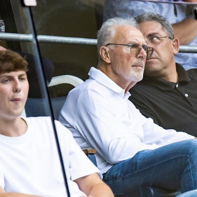 Franz Beckenbauer sitzt im Publikum bei einem Fußballspiel. Neben ihm ein Mann. Beckenbauer trägt eine getönte Brille und schaut mit ernster Miene auf das Spielfeld. Er trägt ein weißes Oberteil.