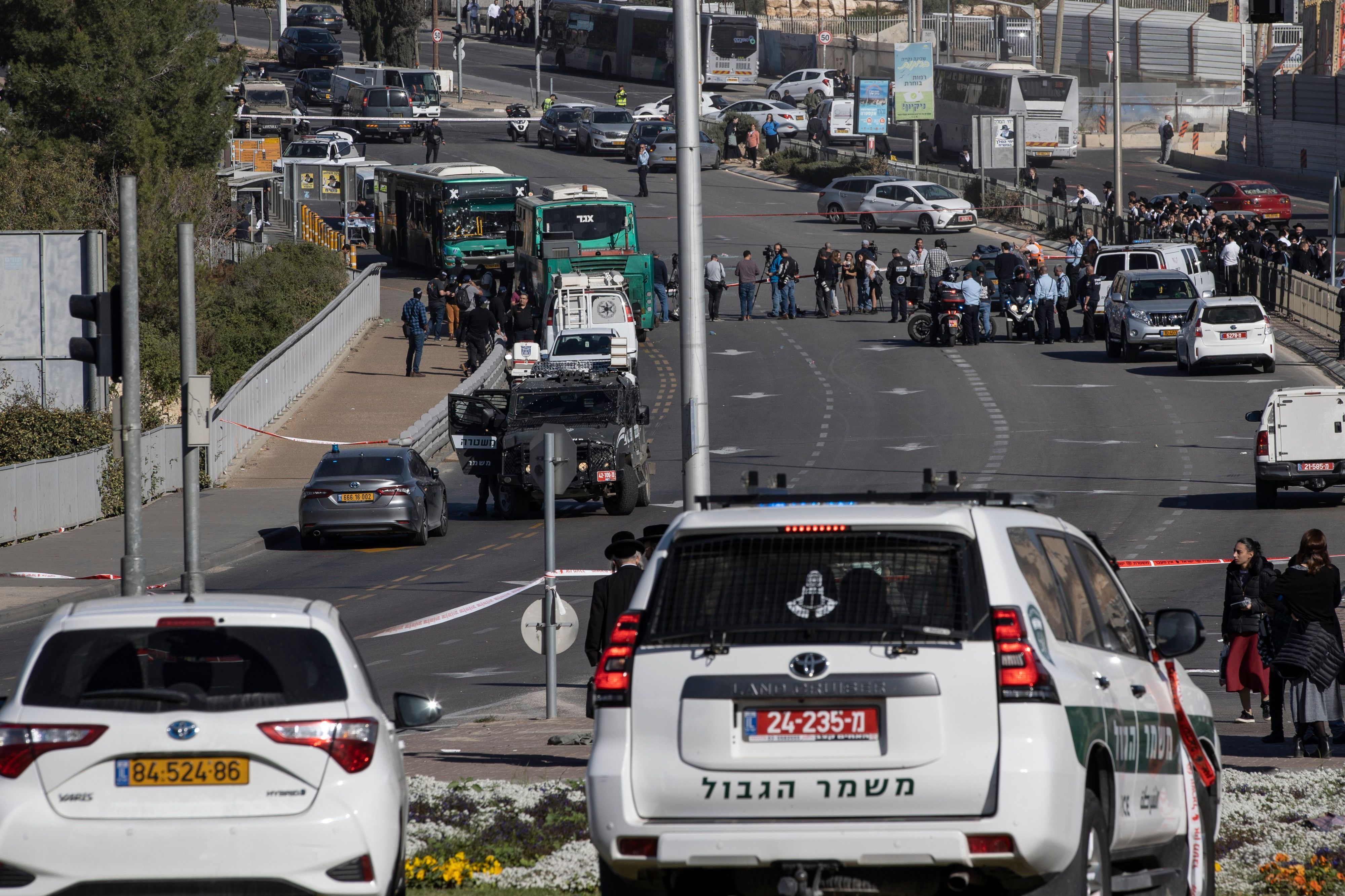 Eine abgesperrte Straße in Jerusalem. Im Hintergrund sind zwei Busse zu sehen, an der Haltestelle kam es zu Explosionen. Polizeiautos, Sicherheitskräfte und Zivilisten stehen auf der Straße.