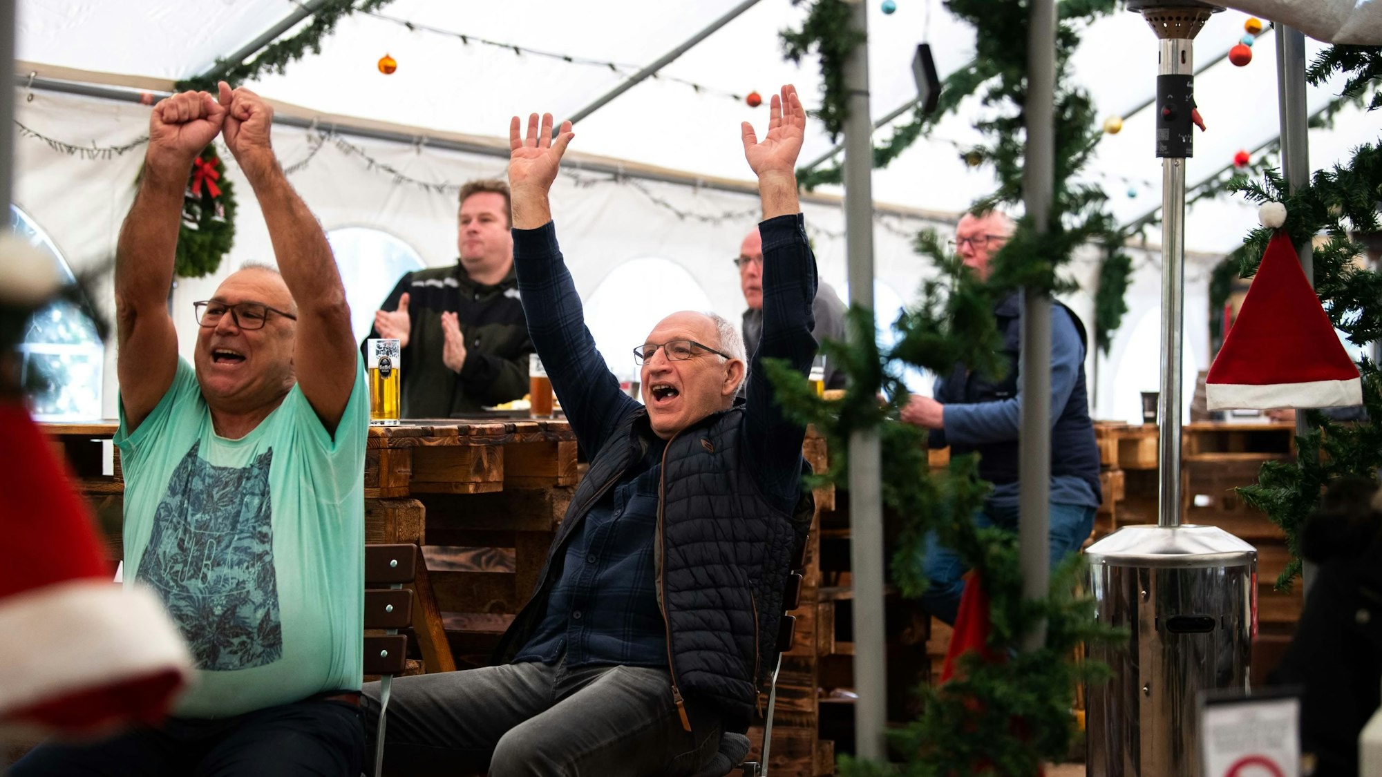 Fußballfans im Biergarten in Kommern werfen jubelnd die Arme in die Höhe und freuen sich über das 1:0 der deutschen Nationalmannschaft gegen Japan. Im Zelt im Biergarten hängt Weihnachtsdekoration.