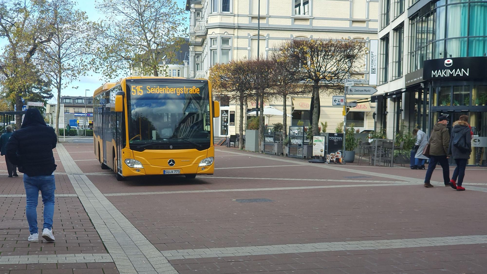 Ein Linienbus der Rhein-Sieg-Verkehrsgesellschaft (RSVG) fährt über eine gepflasterte Straße, die von Bäumen gesäumt ist. Passanten gehen an beiden Seiten.