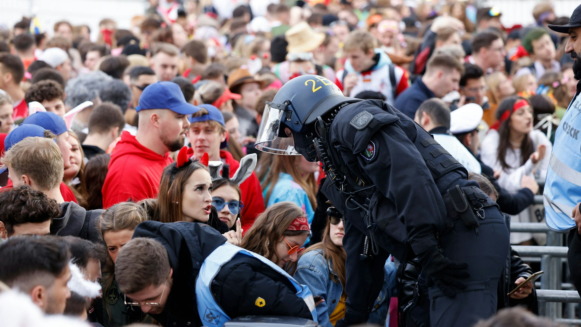 Ein Polizist kommuniziert mit einer jungen Frau im Teufel-Kostüm, im Hintergrund stauen sich Menschenmassen vor den Absperrungen zur Zülpicher Straße.