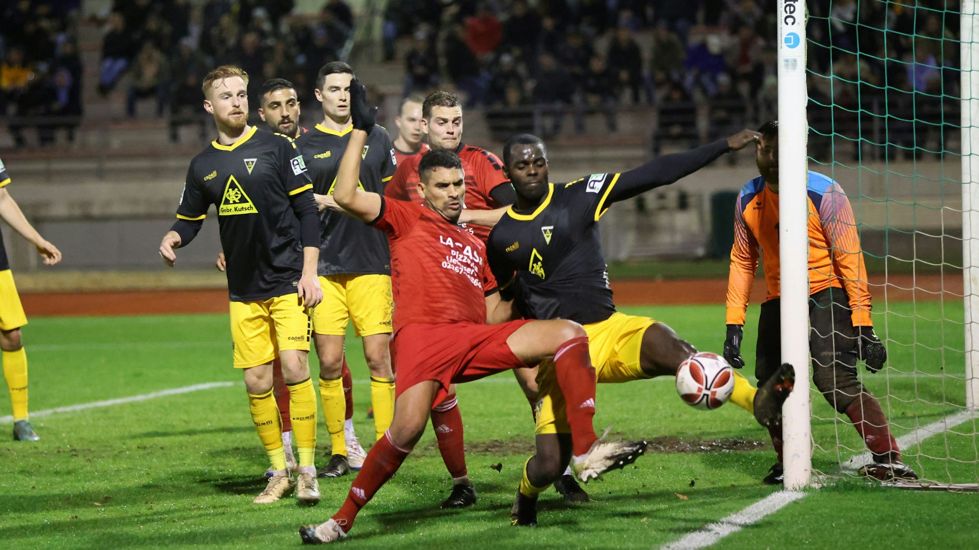 Im Strafraum des VfR Wipperfürth kommt VfR-Spieler Wael Majouj vor dem Aachener Spieler Exauce Andzouana an den Ball und klärt das Spielgerät ins Toraus. Im Hintergrund eine vollbesetzte Tribüne in der Bergisch Gladbacher Belkaw-Arena.
