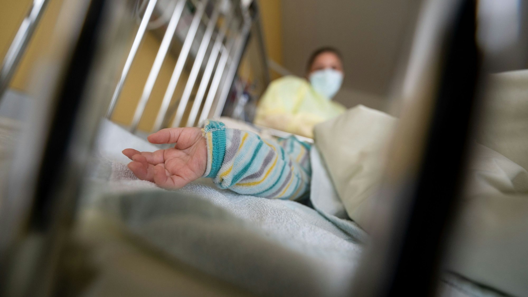 Ein Kind liegt auf einer Kinderstation in einer Klinik. Es sind nur Hand und Arm zu sehen, im Hintergrund steht eine Medizinerin.