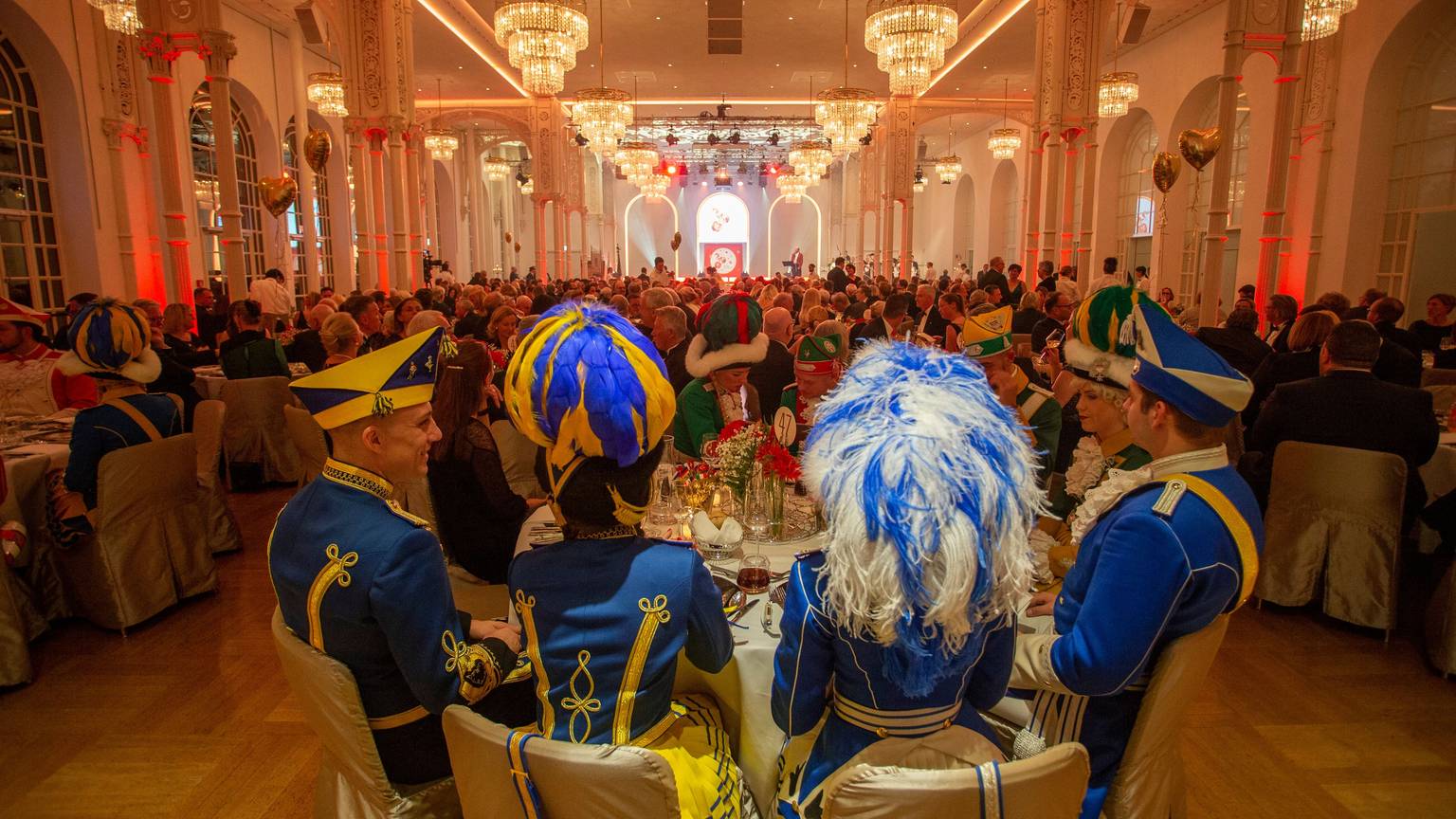 In der prunkvoll gestalteten Flora sitzen rund 500 Gäste an runden Tischen. Im Vordergrund sitzen Mitglieder der Tanzgruppen des Kölner Traditionskorps.