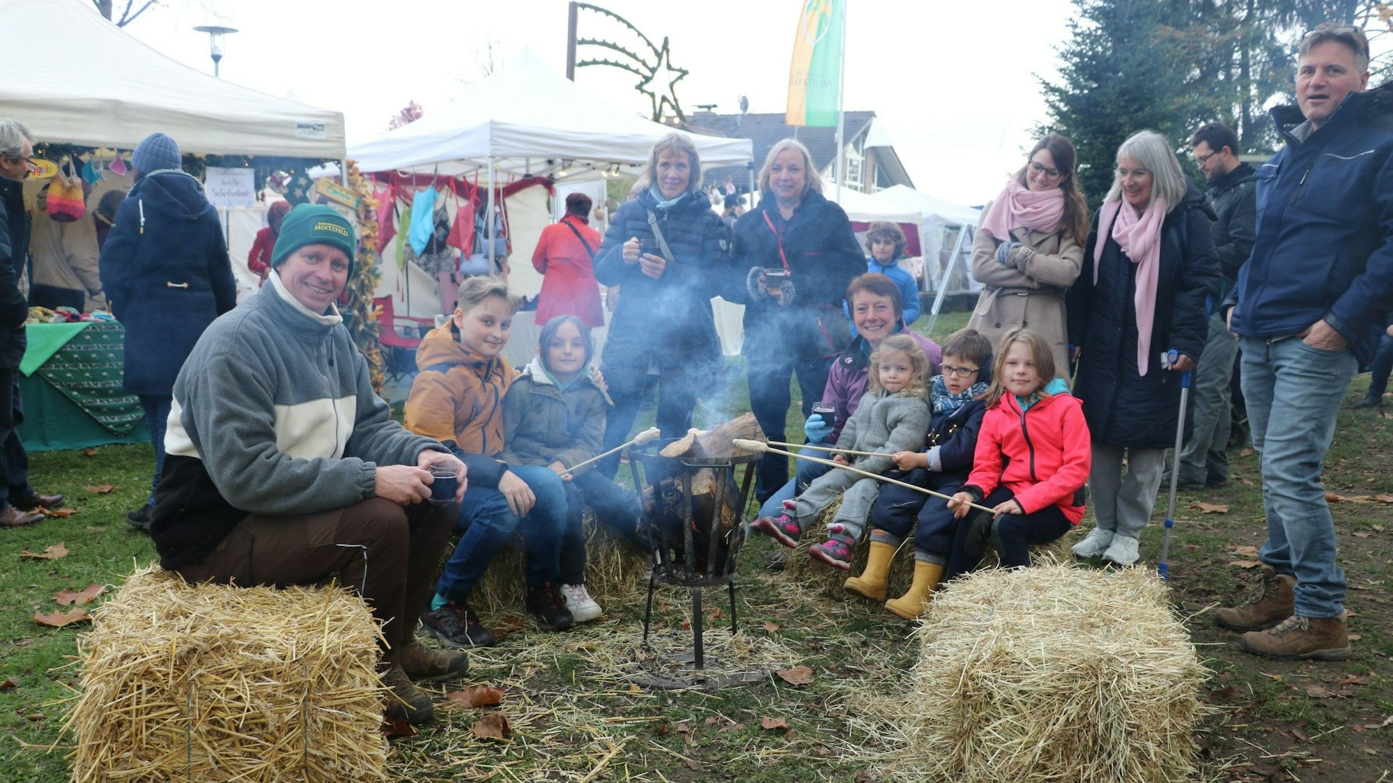 Eine Gruppe aus Erwachsenen und Kindern sitzt auf Strohballen um ein kleines Feuer herum und brät Stockbrot.