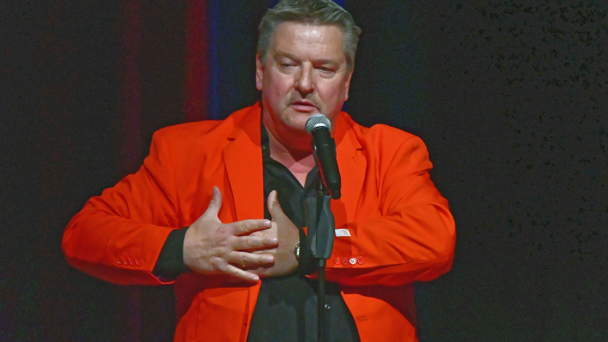 Kurt Kokus steht in schwarzem Hemd und rotem Sacko vor einem dunklen Vorhang in der Alten Drahziherei in Wipperfürth. Vor ihm steht auf einem Ständer ein Funkmikrofon.