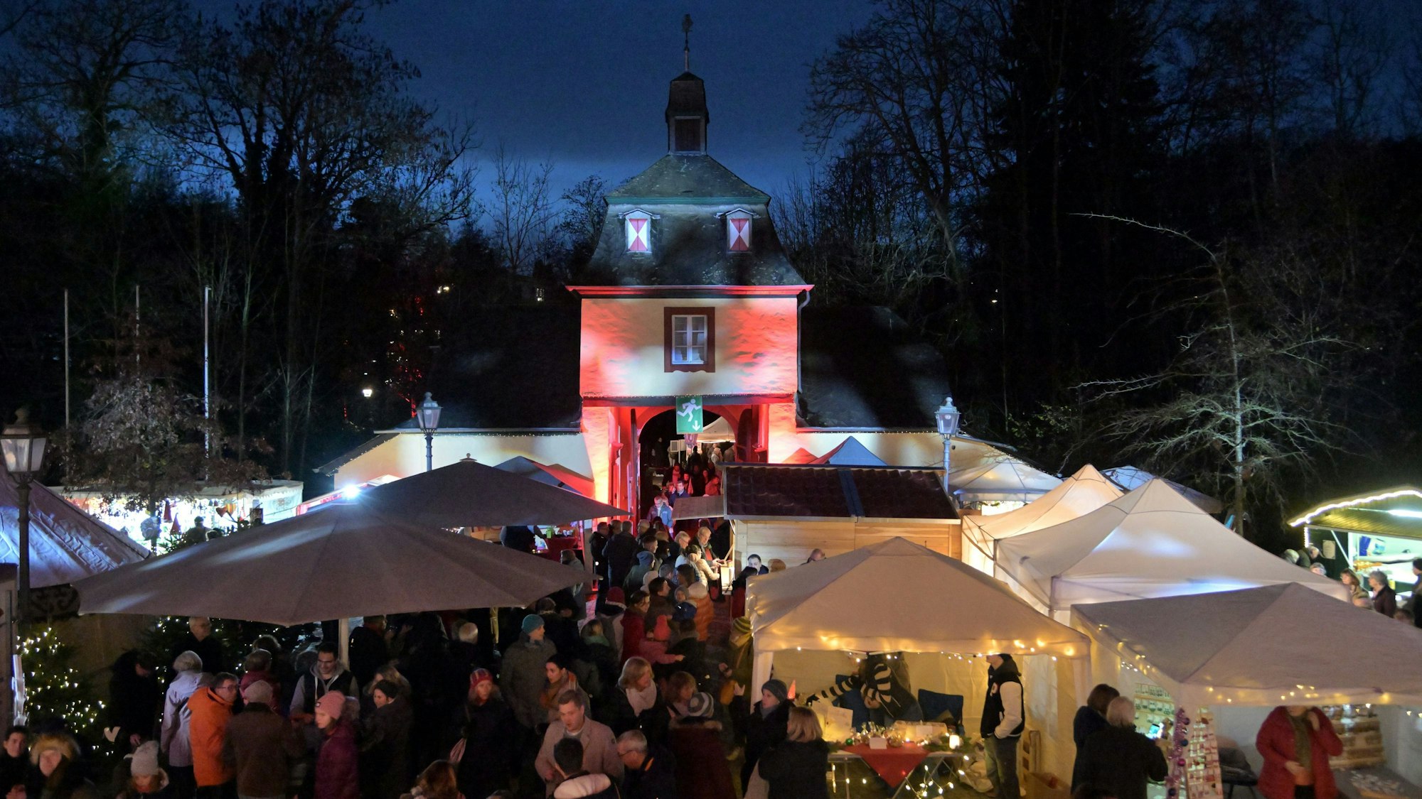 Ein Blick von oben auf den Weihnachtsmarkt an Schloss Eulenbroich in Rösrath. Besucher stehen an den zahlreichen, weißen Ständen. Im Hintergrund ist das angestrahlten Tor zum Schlossgelände zu sehen.