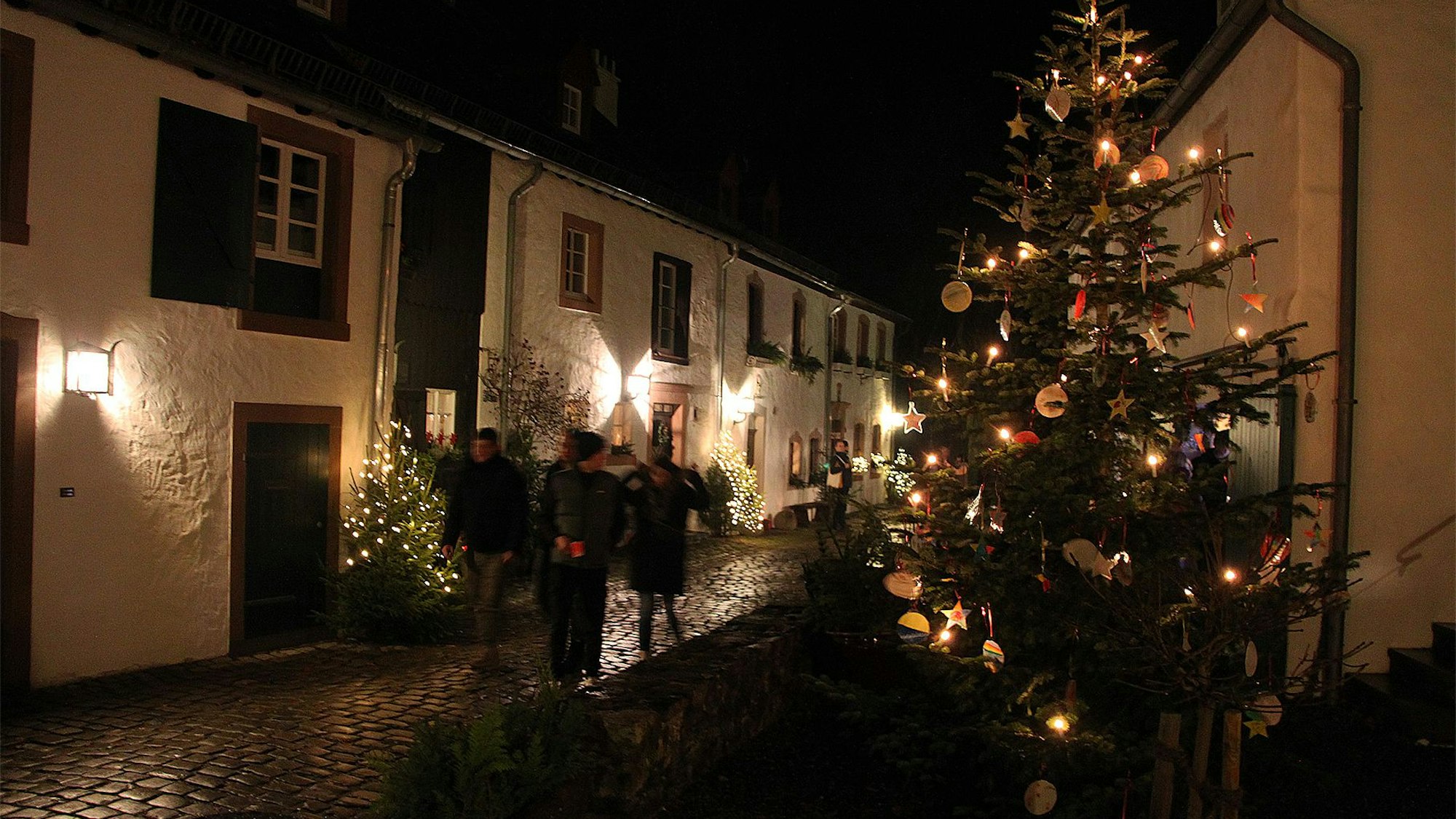 Besucher schlendern an stimmungsvoll geschmückten Weihnachtsbäumen durch die historischen Gassen von Kronenburg.