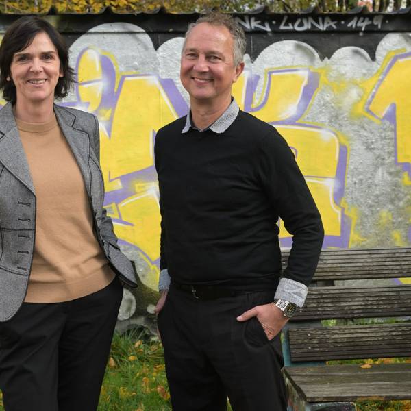 Sigrid Schule und André Eigenbrod, die neuen Leiter der Kreaschule in Bergisch Gladbach, stehen vor einer mit gelb-lilanen Graffitis verzierten Wand.